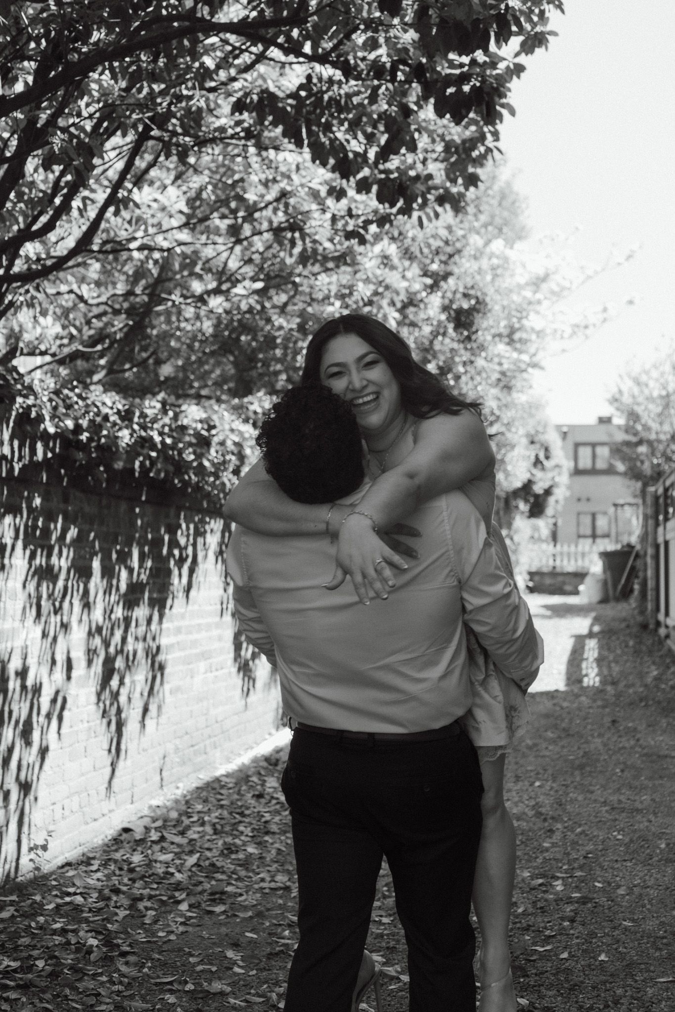 Black and white photo of a smiling woman hugging a man outdoors near trees and a brick wall.