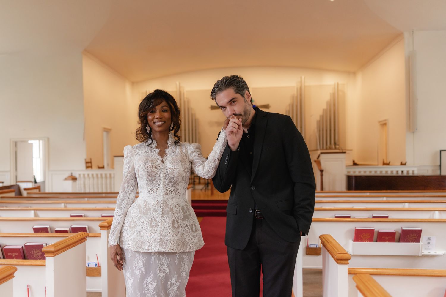Elegant couple posing inside a church, woman in white lace suit, man in black suit, with wooden pews and organ pipes. just eloped.