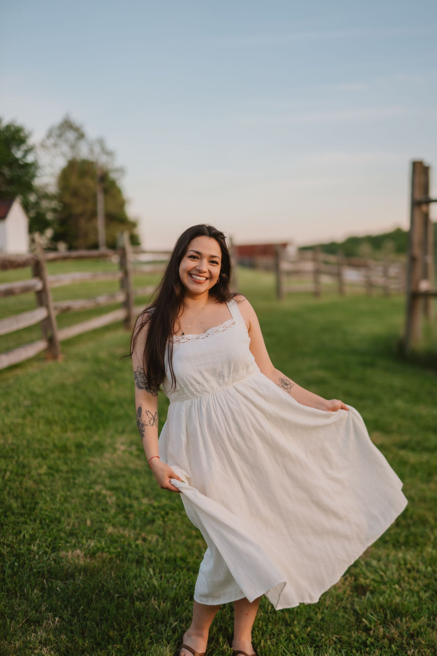 Gift of Moments owner, Tatiana, in a white sundress twirls on a green farm lawn at sunset, showing off her tattoo sleeve.