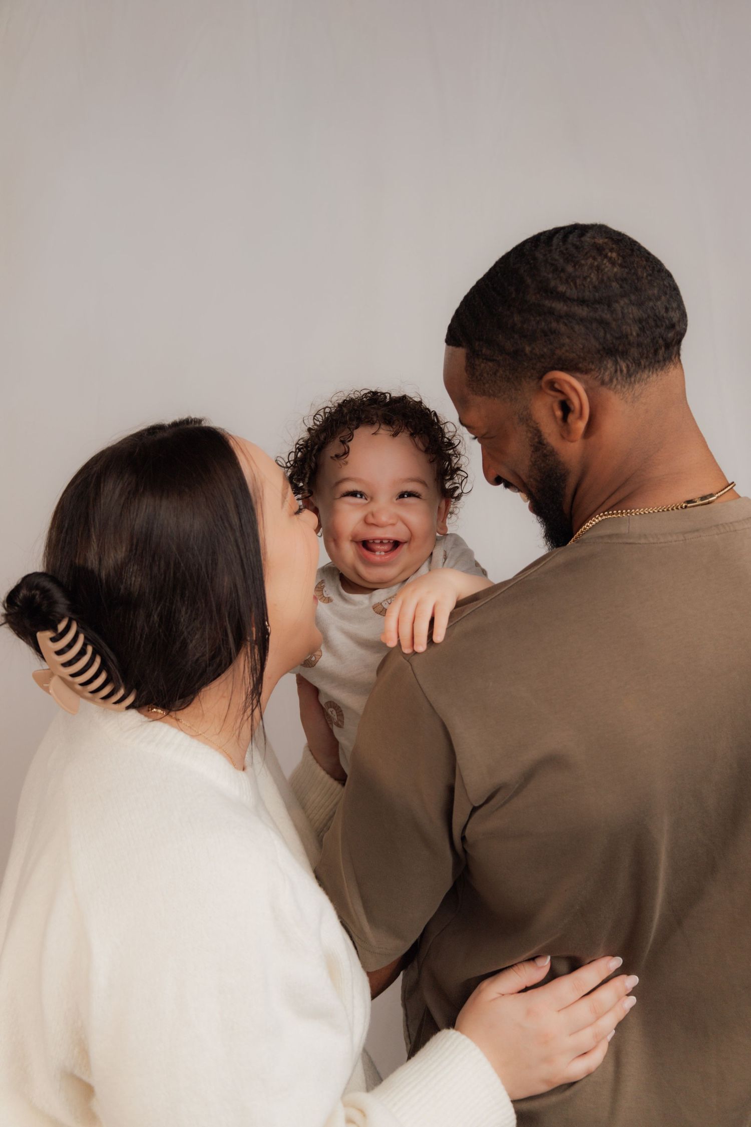 Happy baby laughing with parents in a warm family portrait against a neutral background.
