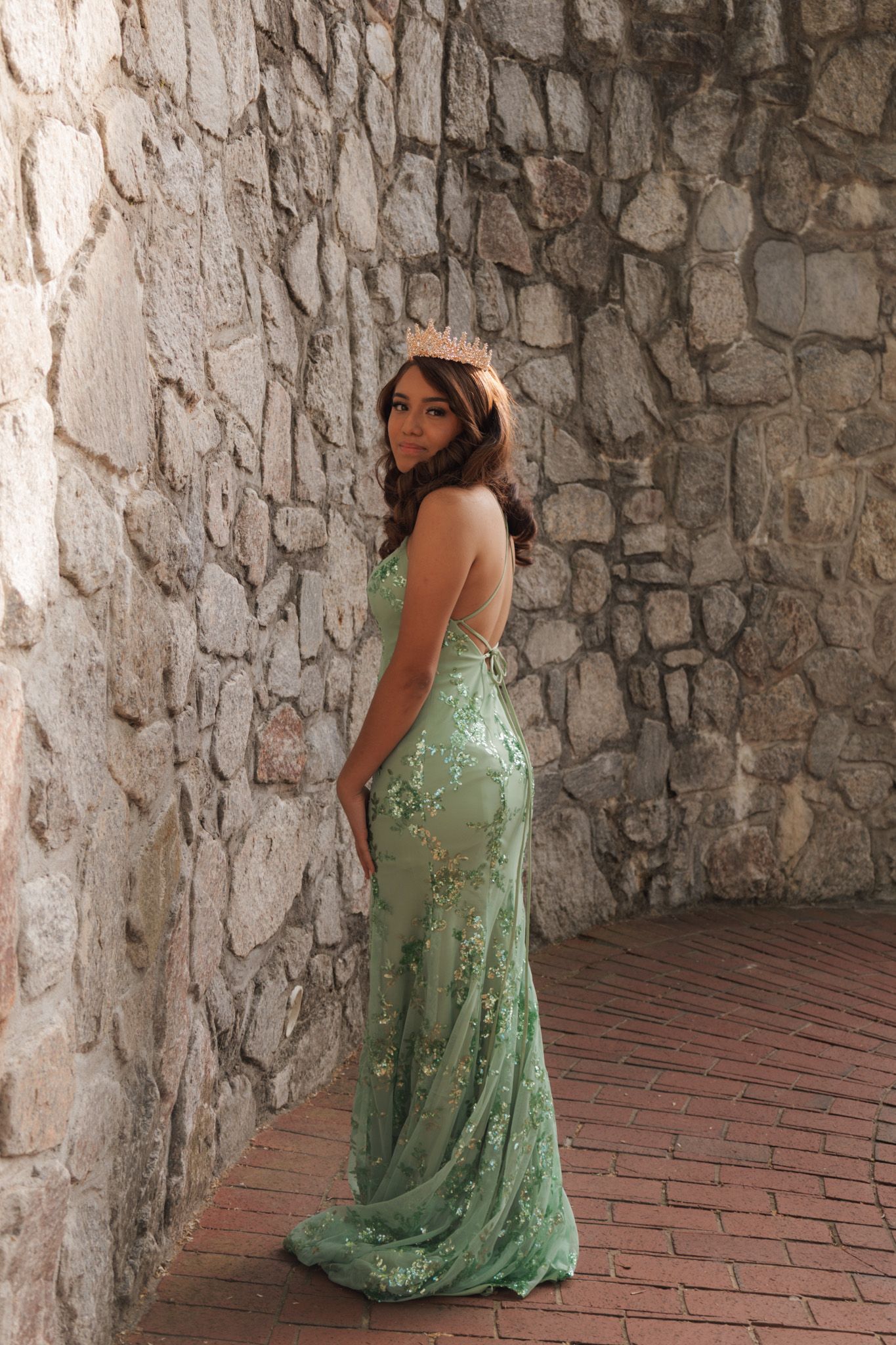Woman in elegant green floral sequin gown posing gracefully against a rustic stone wall on brick flooring.