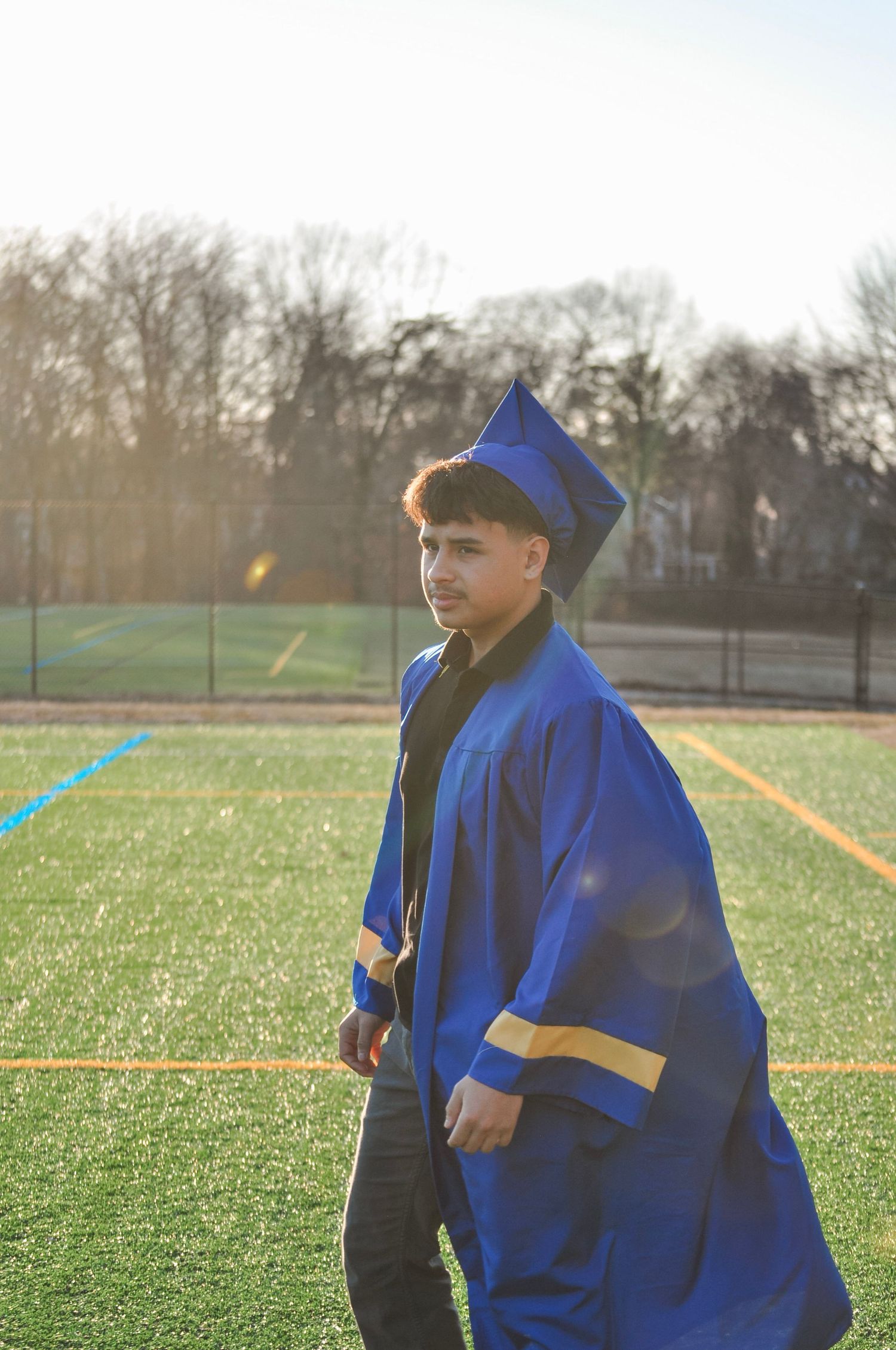 Young man in blue graduation cap and gown standing on a sports field during golden hour sunlight.
