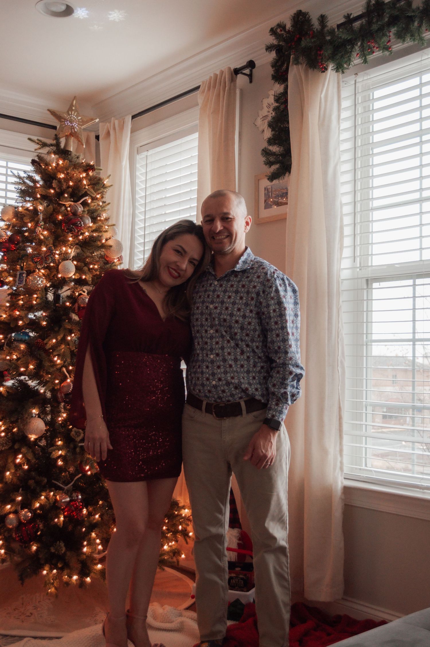 Couple smiling together in front of a decorated Christmas tree, woman in red sequin dress, man in patterned blue shirt.