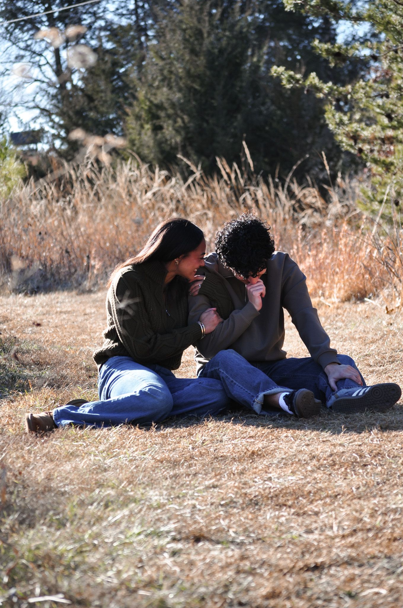 Couple sitting together on dry grass field in autumn, sharing an intimate moment outdoors near tall golden grasses.