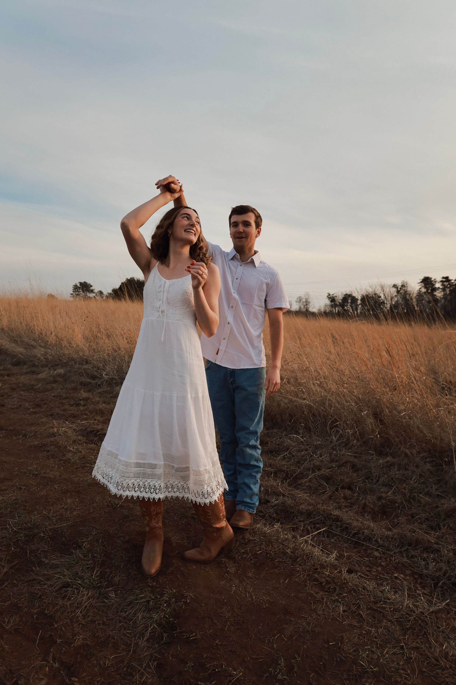 A couple stands in a golden field at sunset, woman in white dress and boots, man in white shirt and jeans.