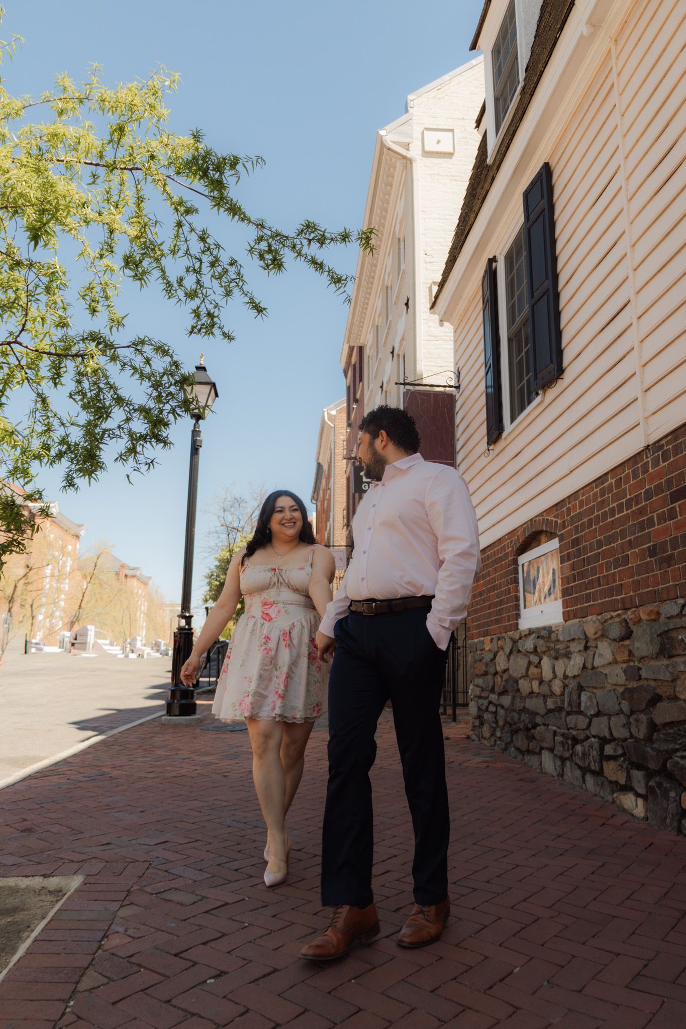 Couple holding hands walking on brick sidewalk in historic town, woman in floral dress, man in pink shirt on sunny day.