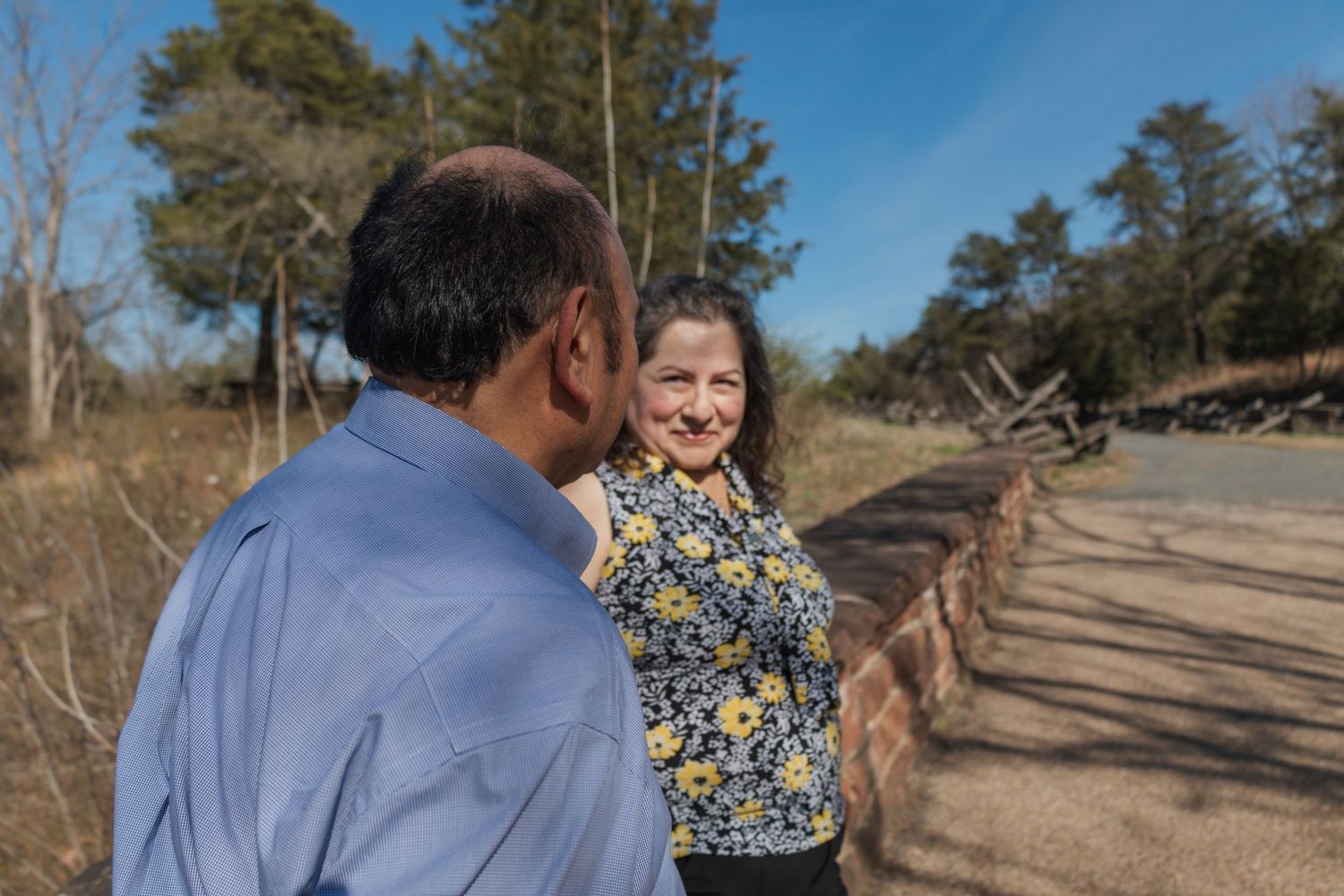 Two people conversing outdoors on a sunny day near a rural road surrounded by trees and open land.