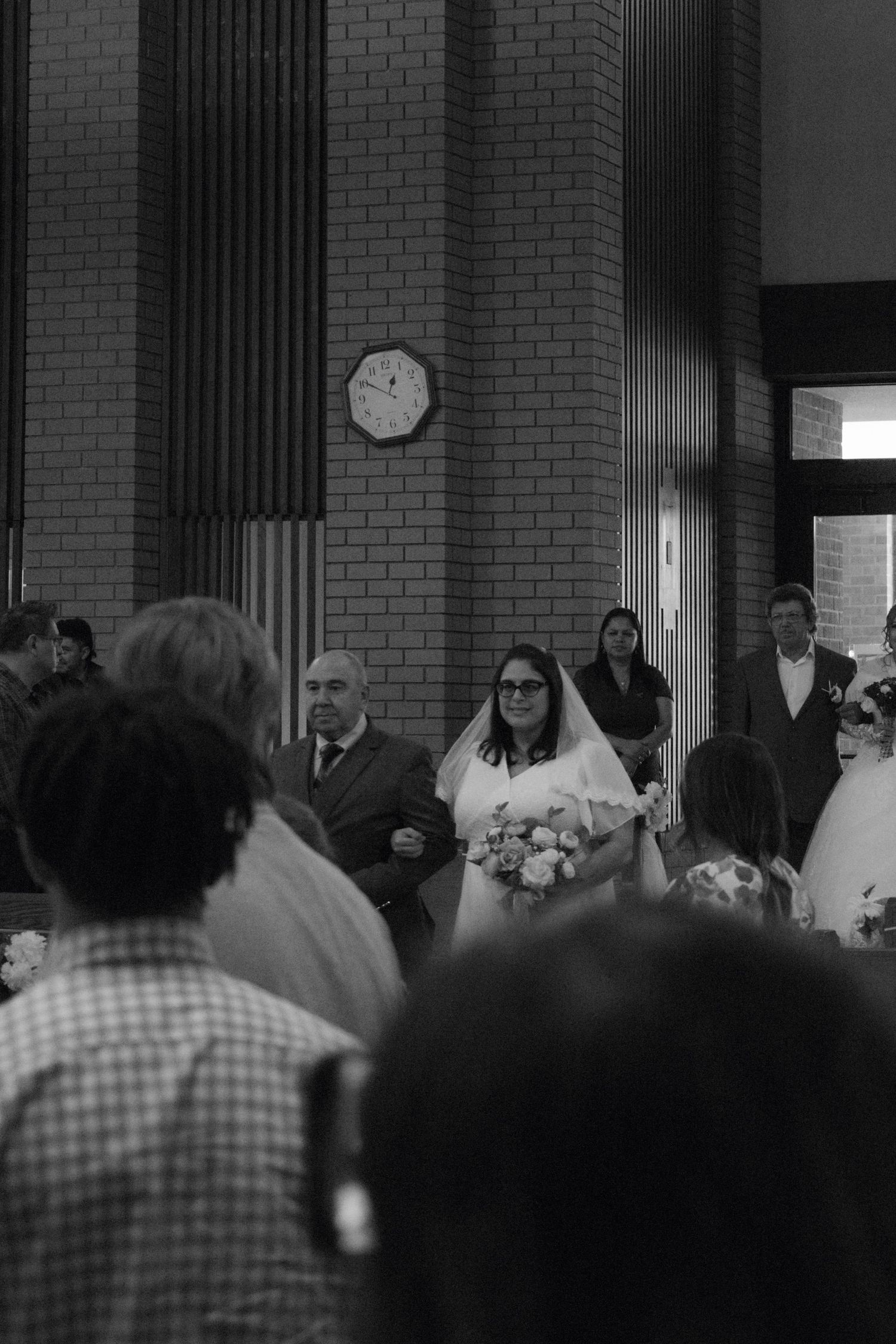 Black and white wedding photo of a bride in white gown holding bouquet walking down church aisle with guests.