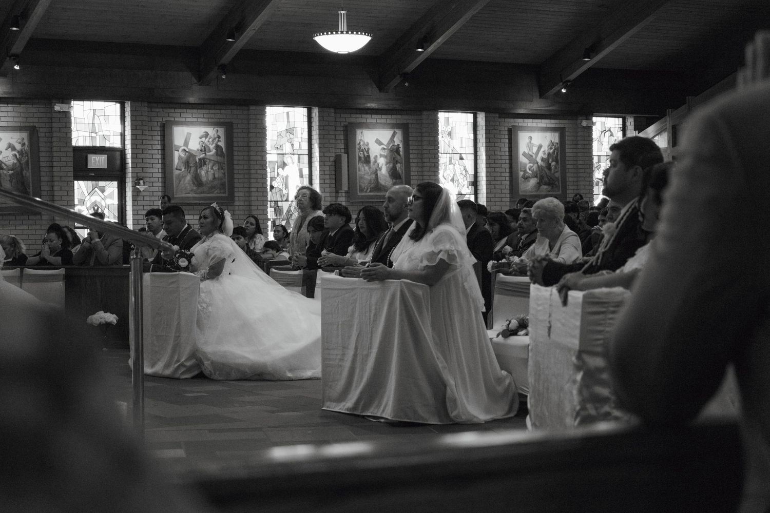 Black and white photo of a bride in a white gown kneeling at church pews during a traditional wedding ceremony.