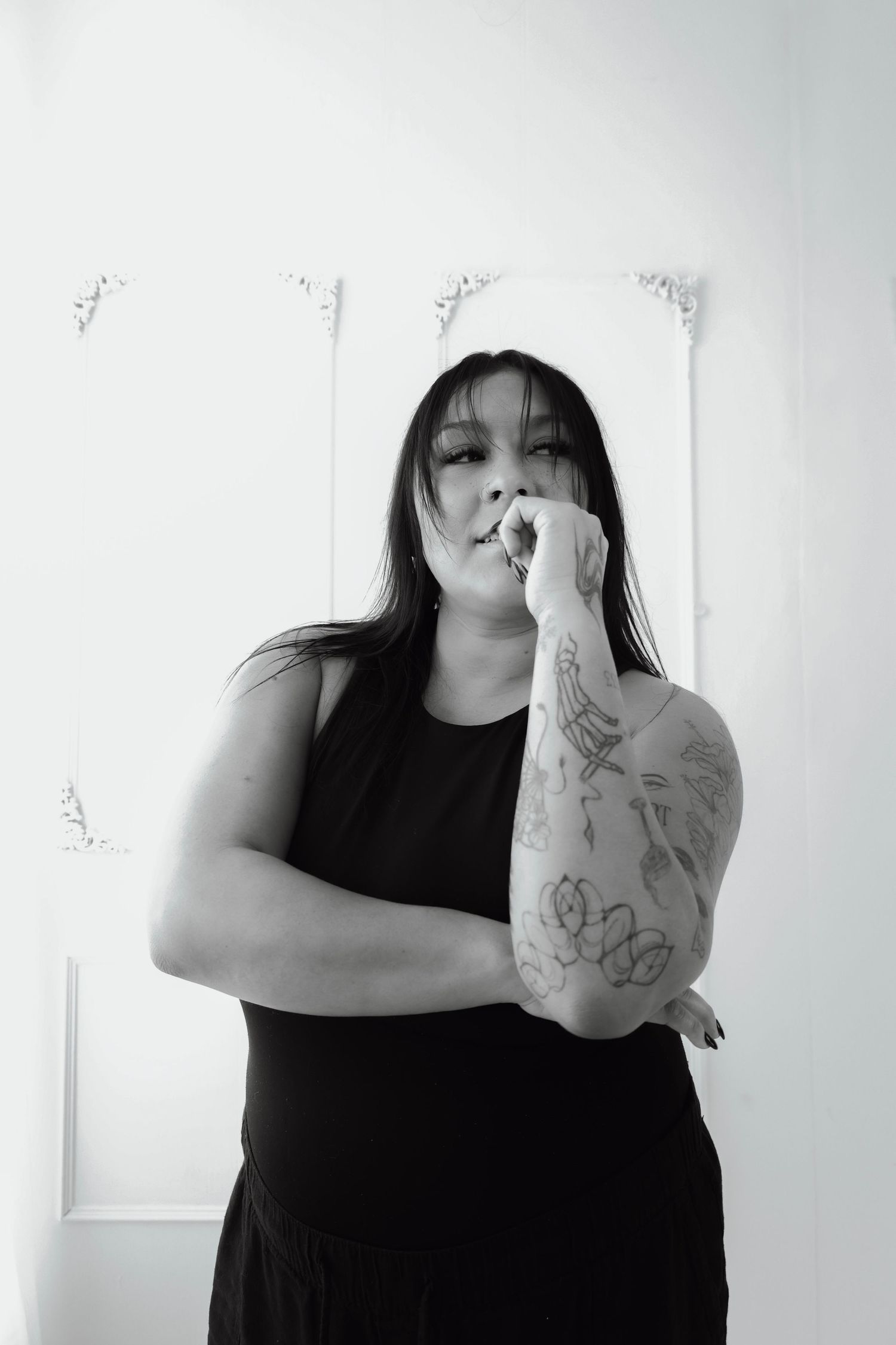 Black and white portrait of a woman in a black dress with tattoos on her arm, posing thoughtfully in a studio.