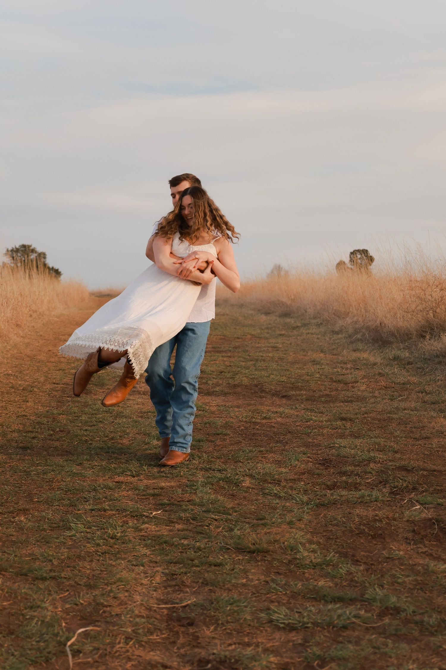 Man in jeans spinning woman in white dress on a rural path at golden hour.