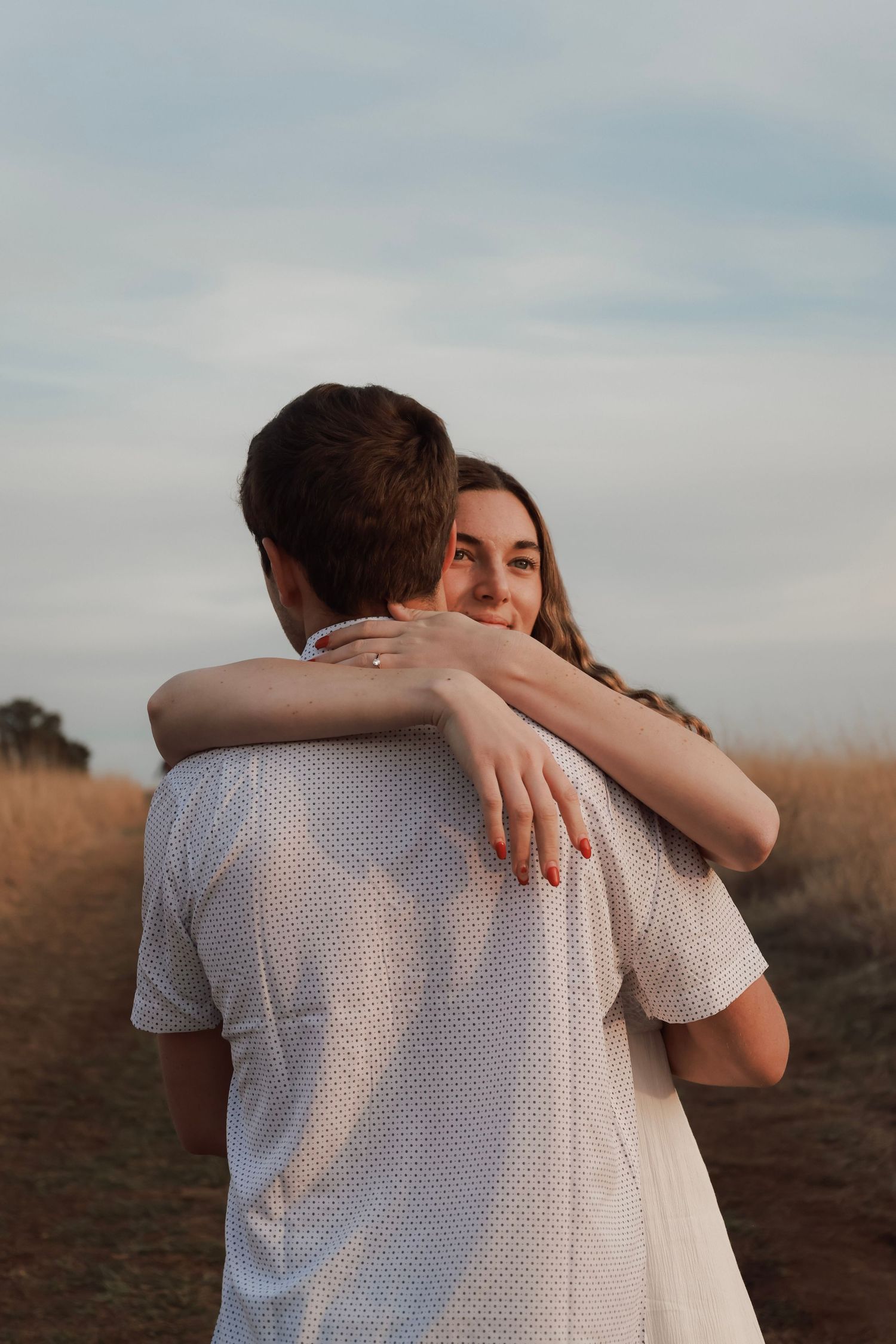 A couple embraces in a golden field at dusk, woman smiling over mans shoulder wearing white outfits.