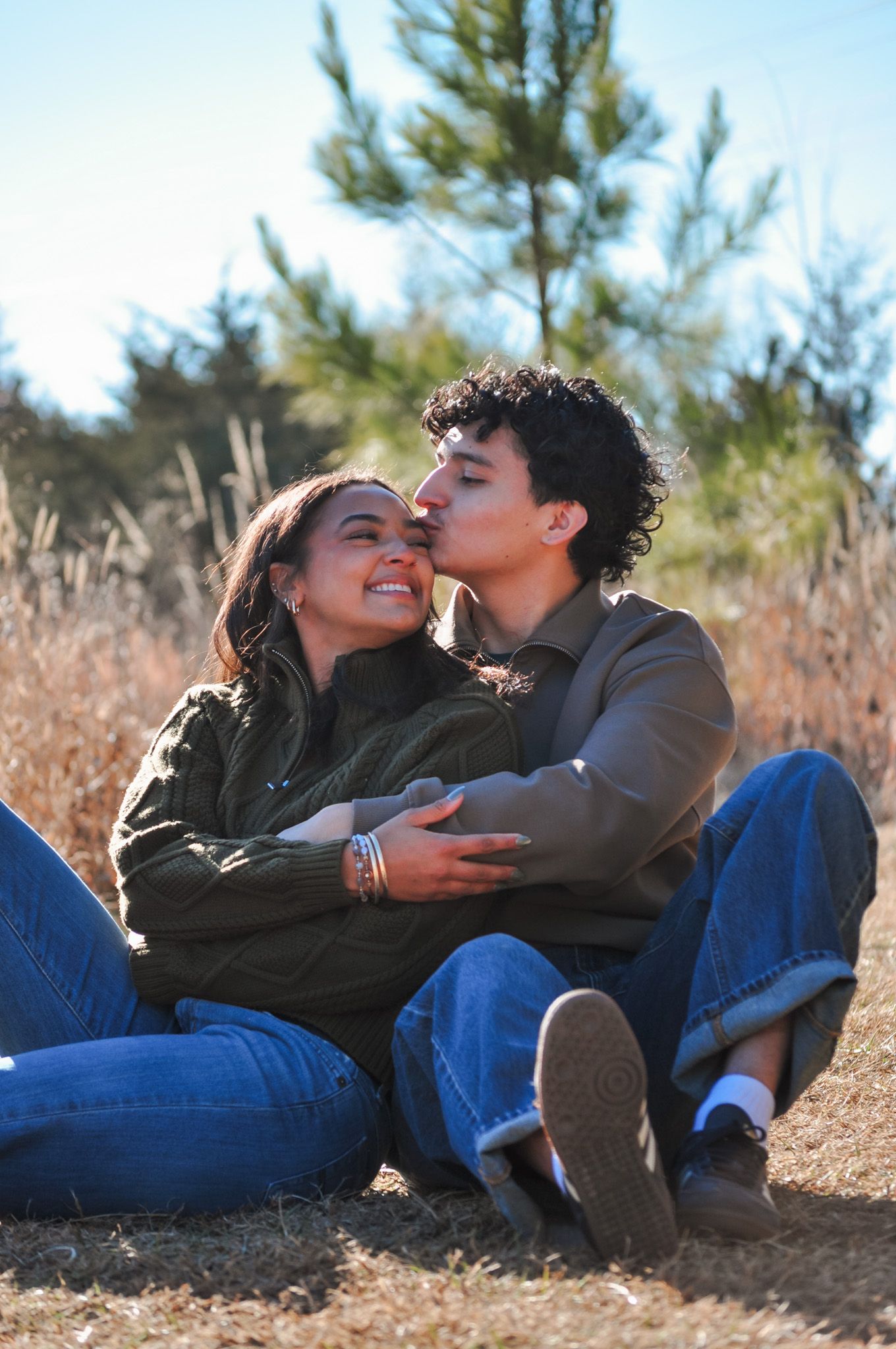 Young couple sitting outdoors in a sunny field, sharing a tender kiss while embracing in casual denim outfits.