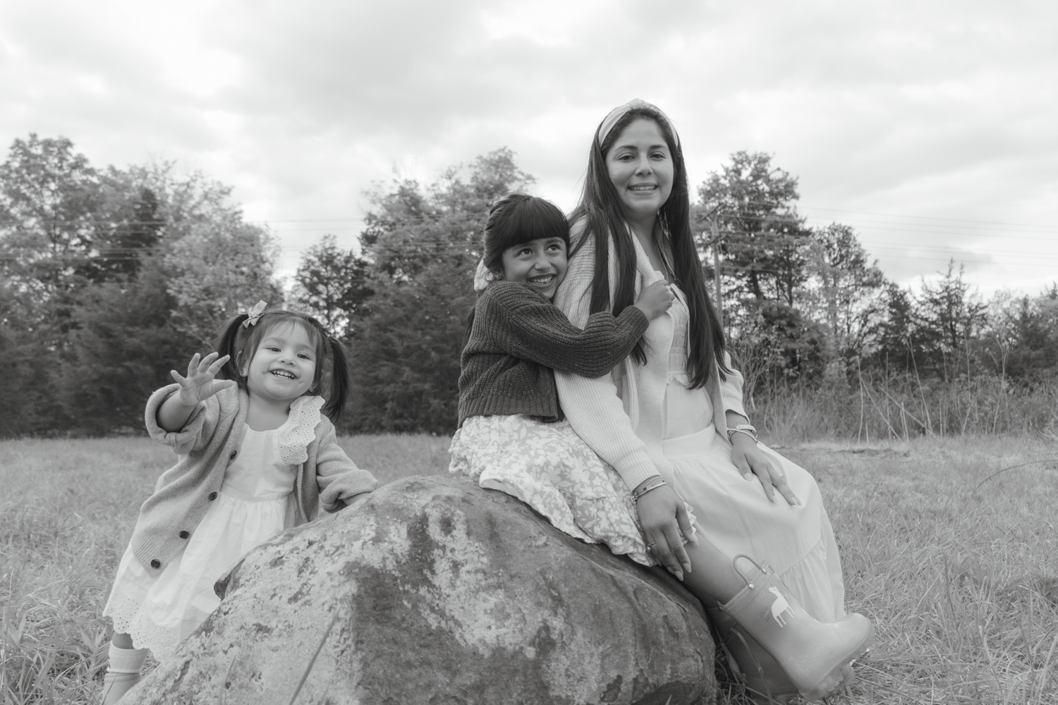 Black and white photo of three children sitting on a rock in a field with trees in the background.