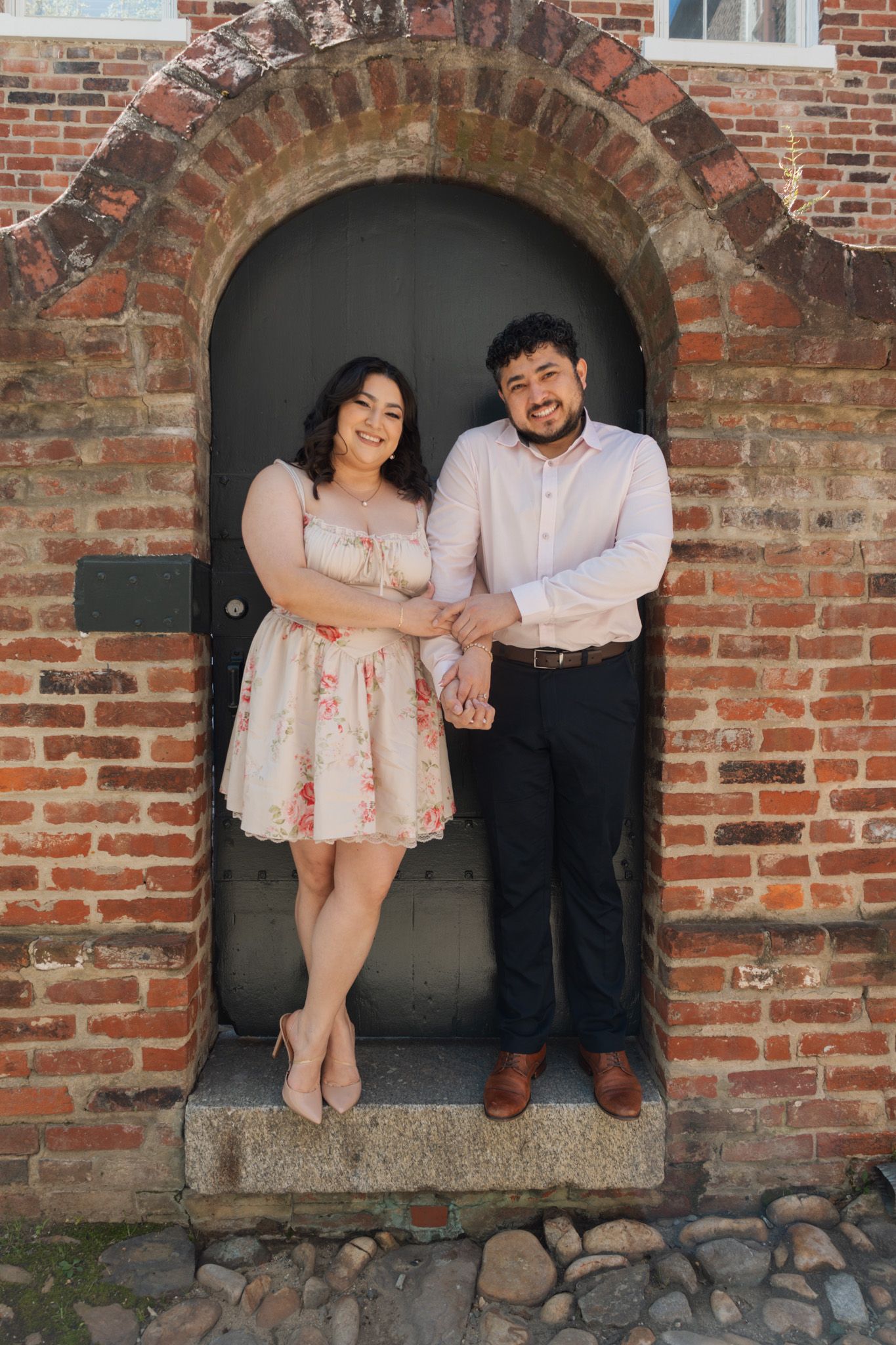 Couple holding hands and smiling in front of a brick archway door, woman in floral dress, man in white shirt.