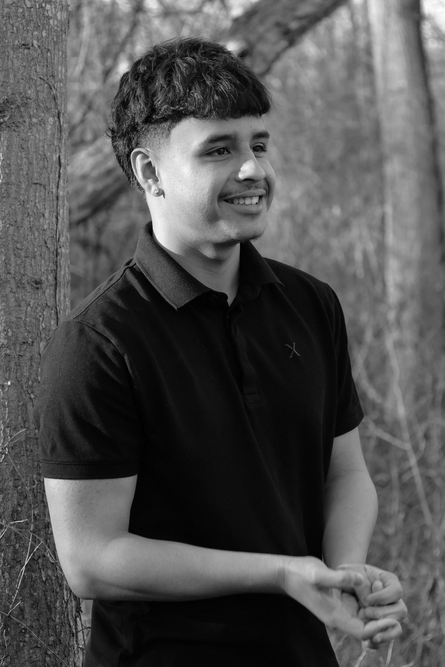Young man smiling in black polo shirt leaning against tree in forest, black and white portrait photo.