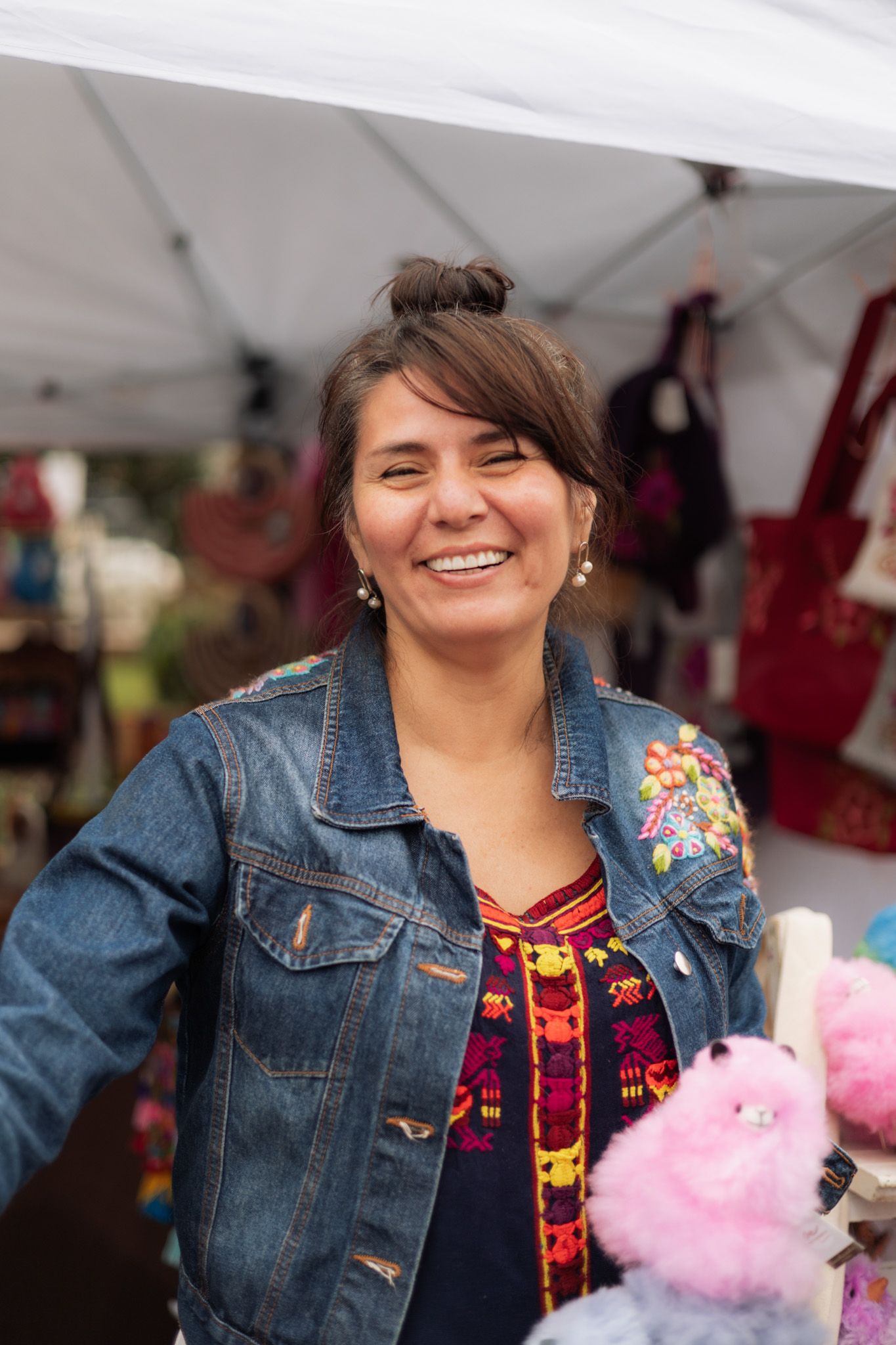 Smiling woman in denim jacket with floral embroidery at outdoor market stall with colorful crafts.