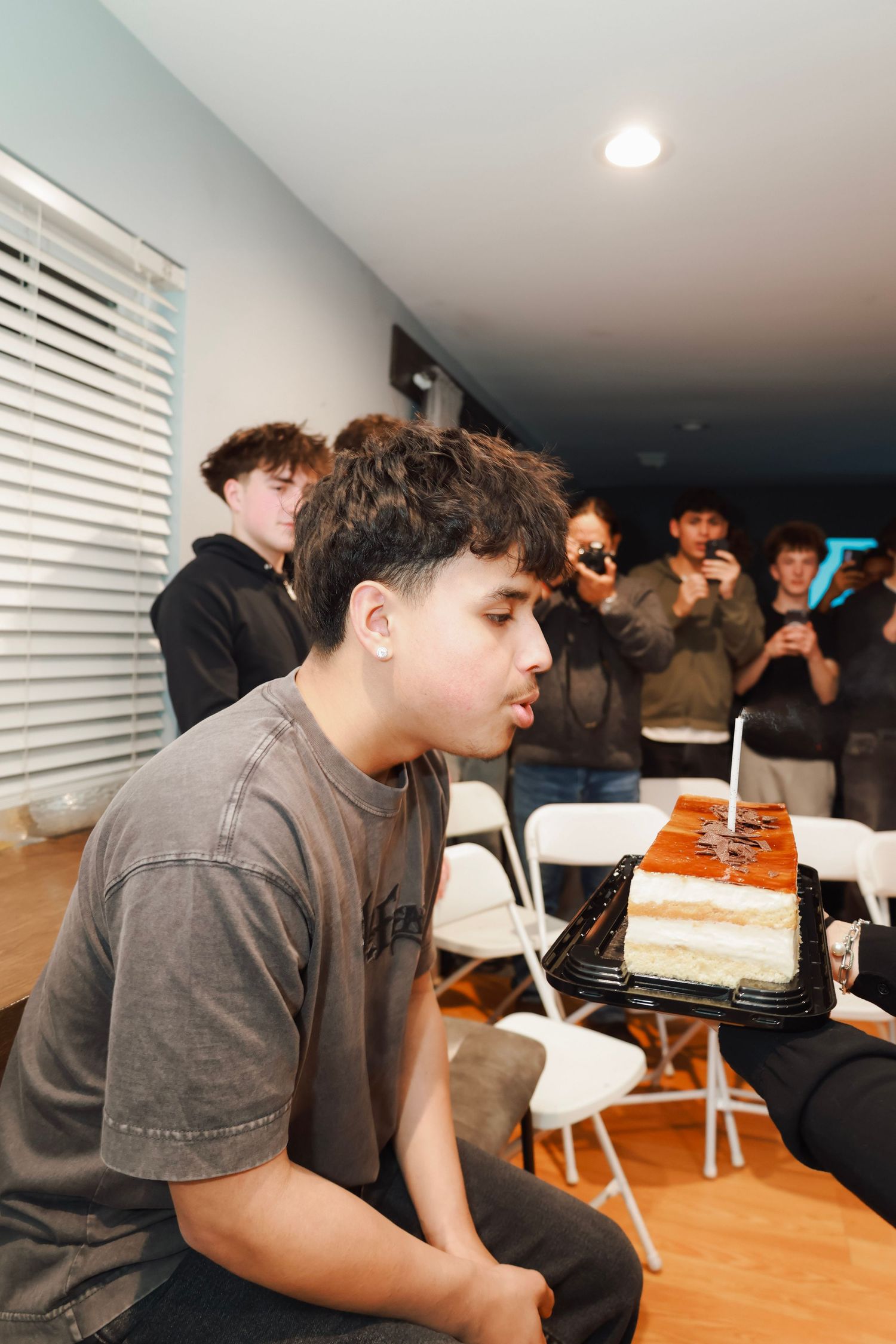 Teen blowing out candle on birthday cake at party while friends photograph the moment.