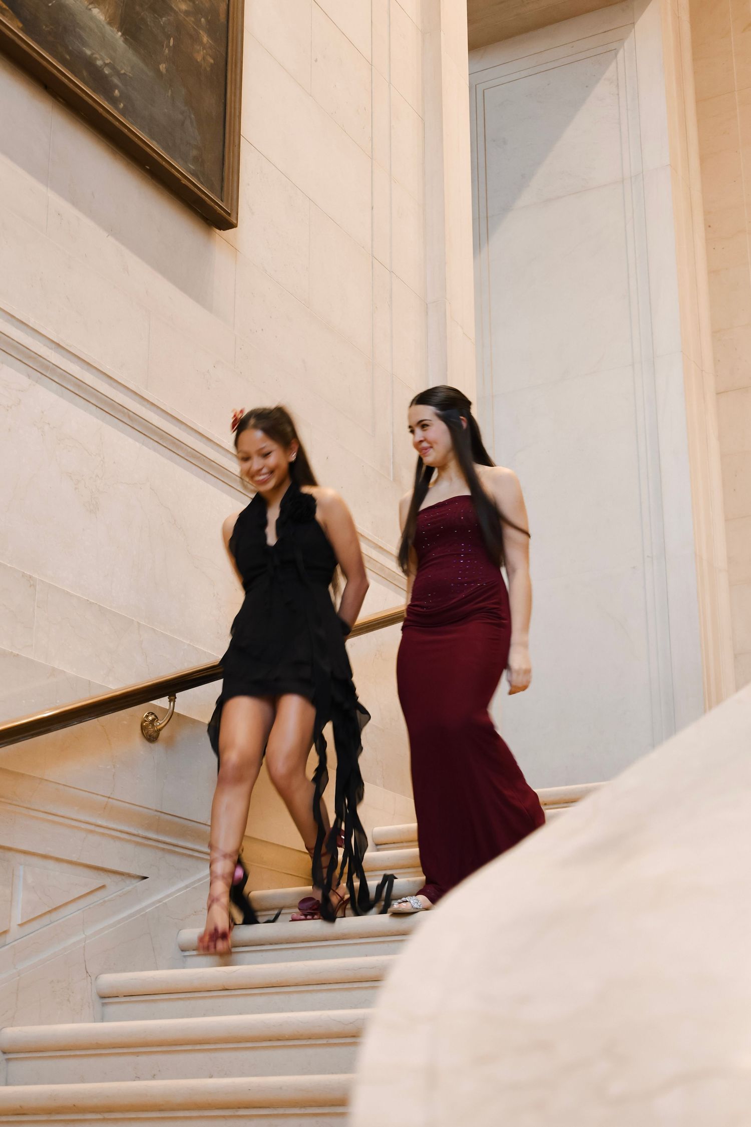 Two elegant women descend marble stairs, one in black ruffled dress, other in burgundy gown, at formal event.