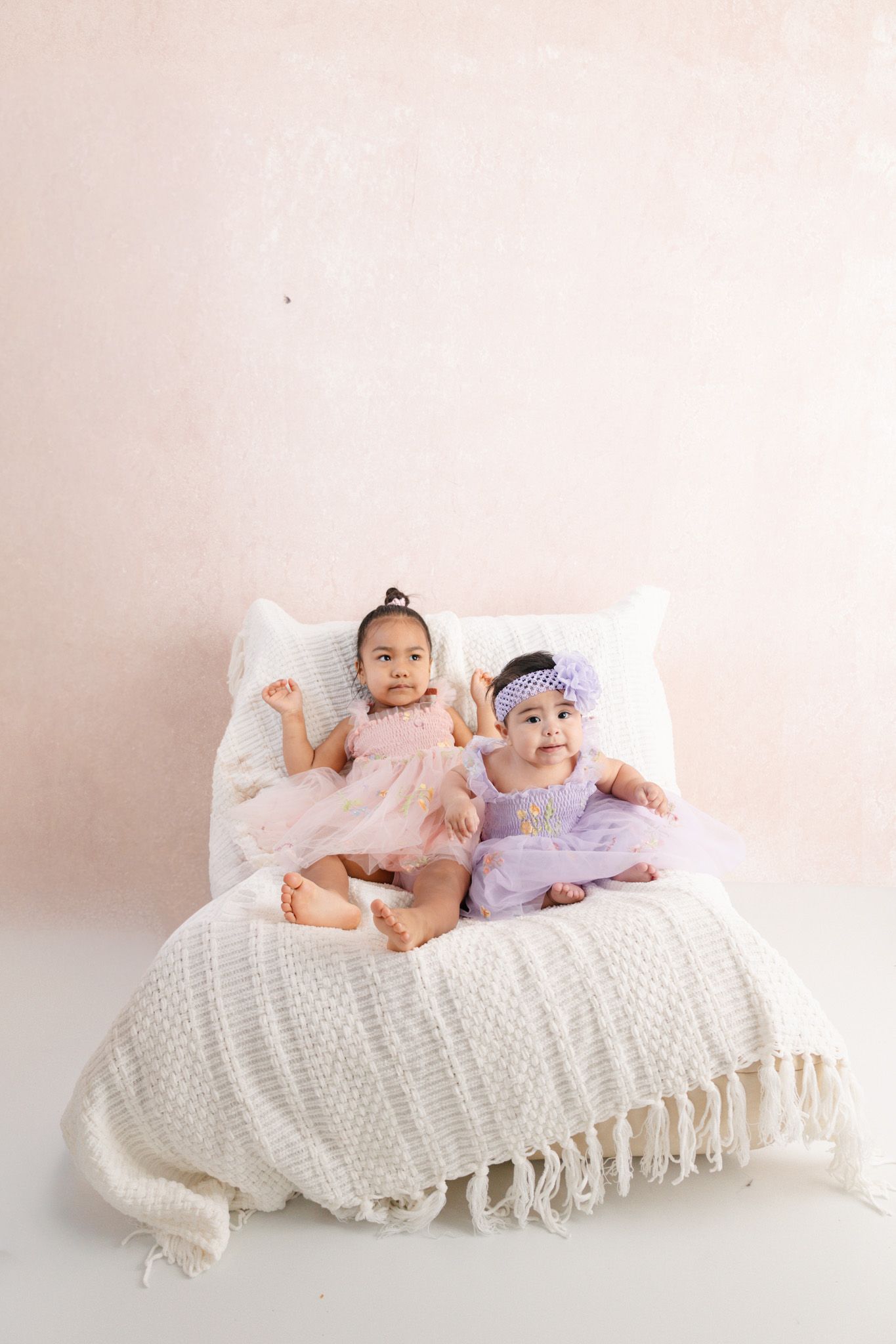 Two baby girls in pink and lavender dresses sitting together on a white bed with fringe blanket against a blush wall.