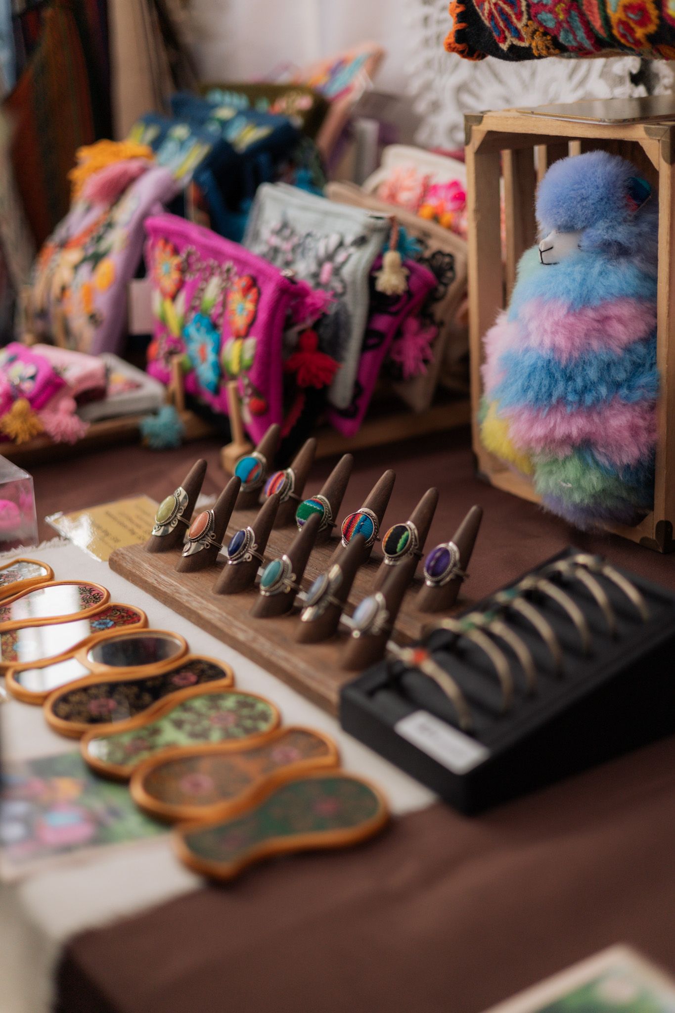 Colorful artisan market display with gemstone rings, embroidered bags, wooden coasters, and a fluffy rainbow alpaca toy.