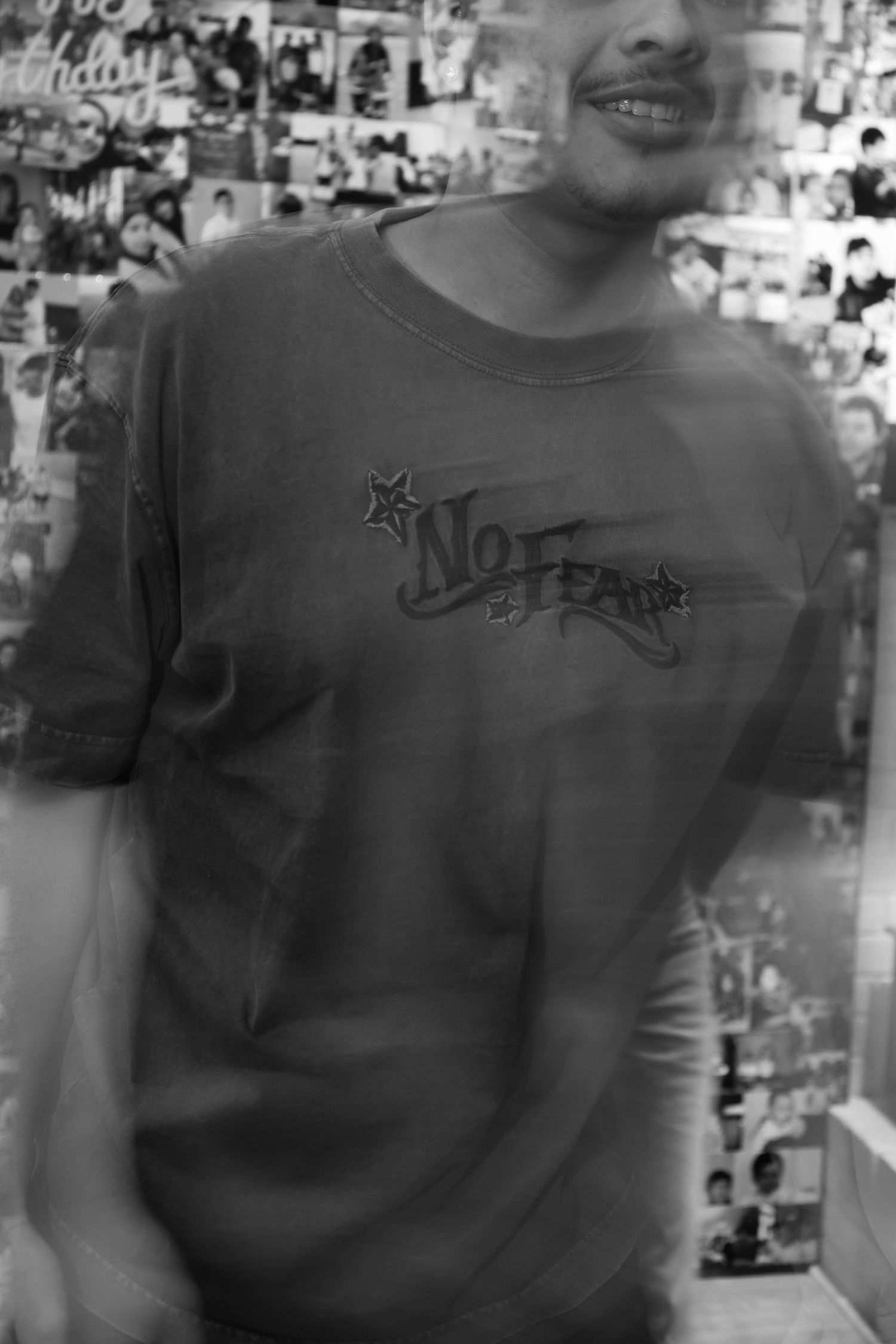 Black and white photo of a smiling person wearing a No Fear graphic t-shirt, with photos covering the wall behind them.