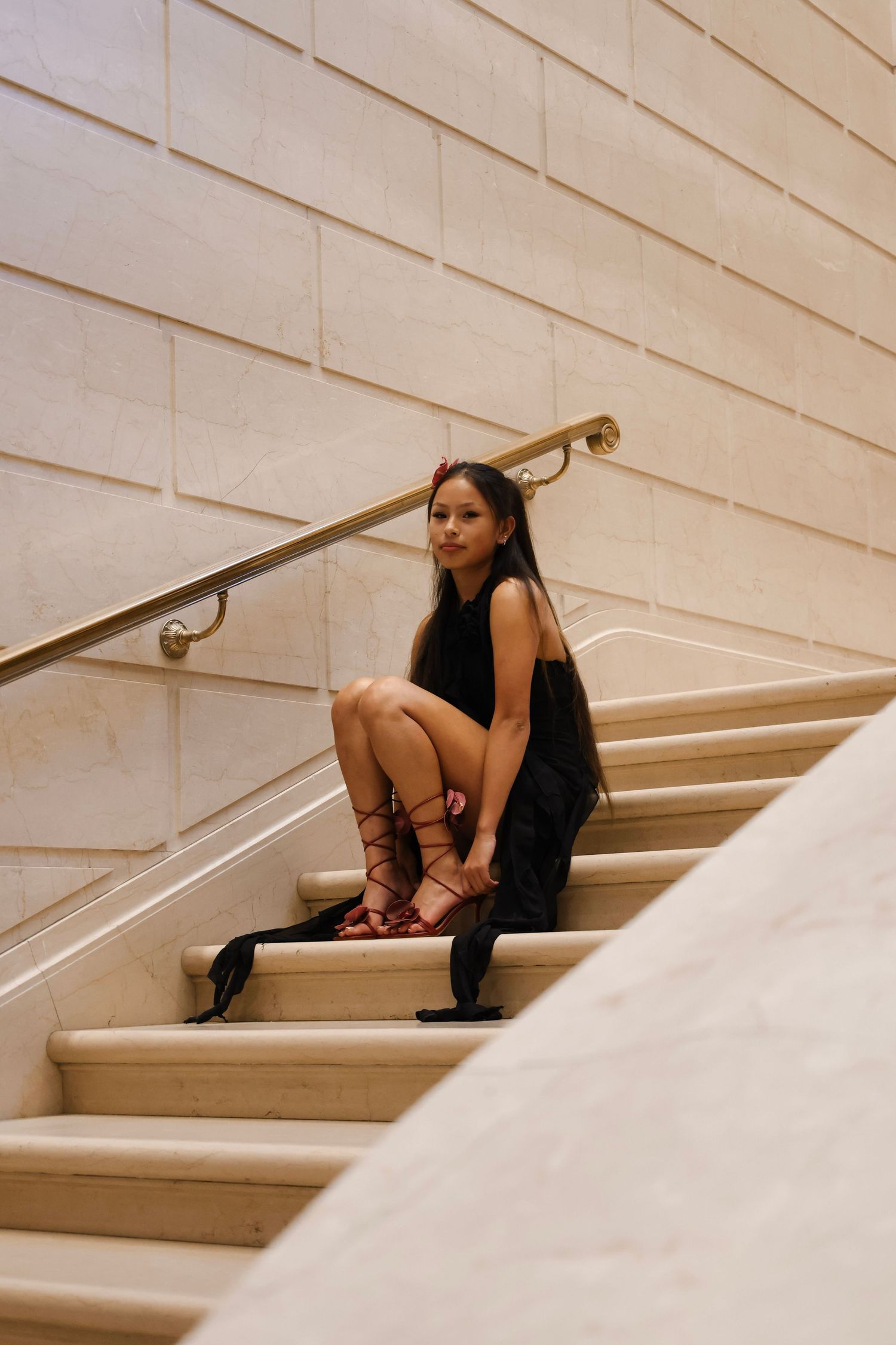 Woman in black dress and red lace-up heels sitting gracefully on marble stairs with gold railing.