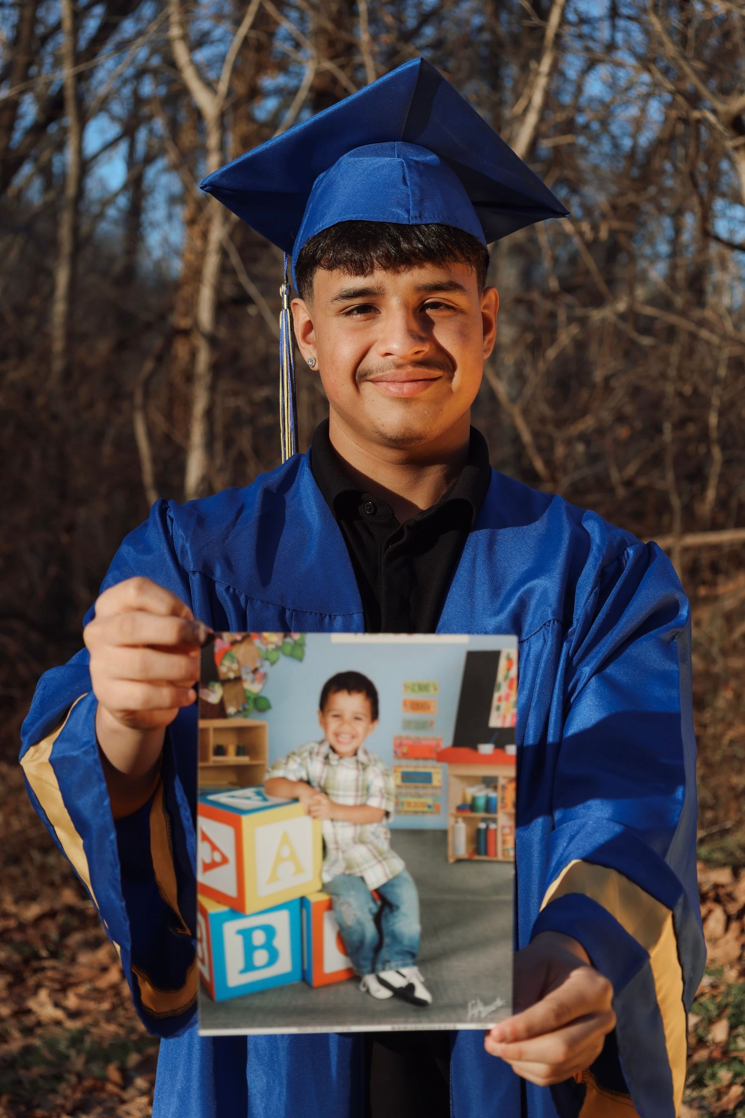 Young man in blue graduation cap and gown proudly holds his kindergarten school photo, smiling outdoors.