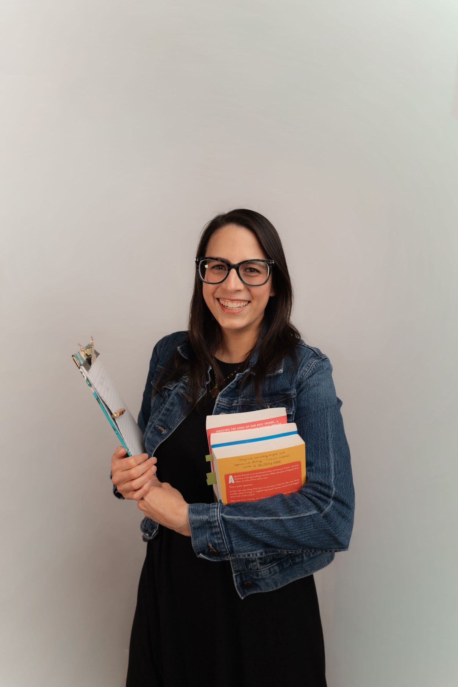 Smiling woman with glasses holding colorful books and a selfie stick, wearing a denim jacket over a black dress.