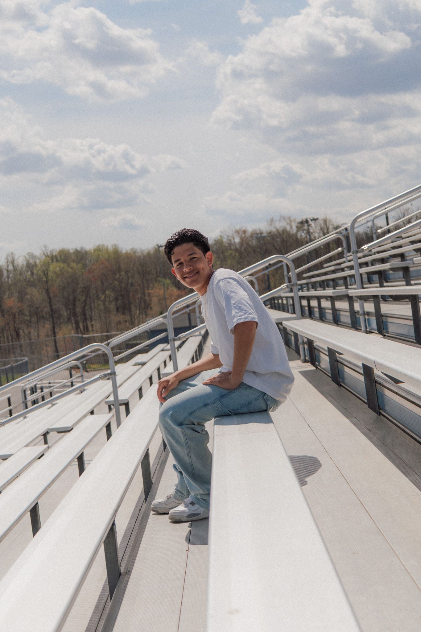 Smiling young man sitting on bleachers in a white t-shirt and jeans on a sunny day.