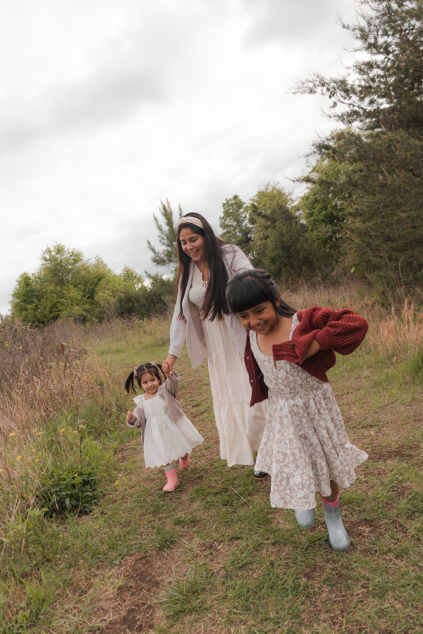 A mother and two daughters walk through a grassy field, laughing and holding hands on a cloudy day.