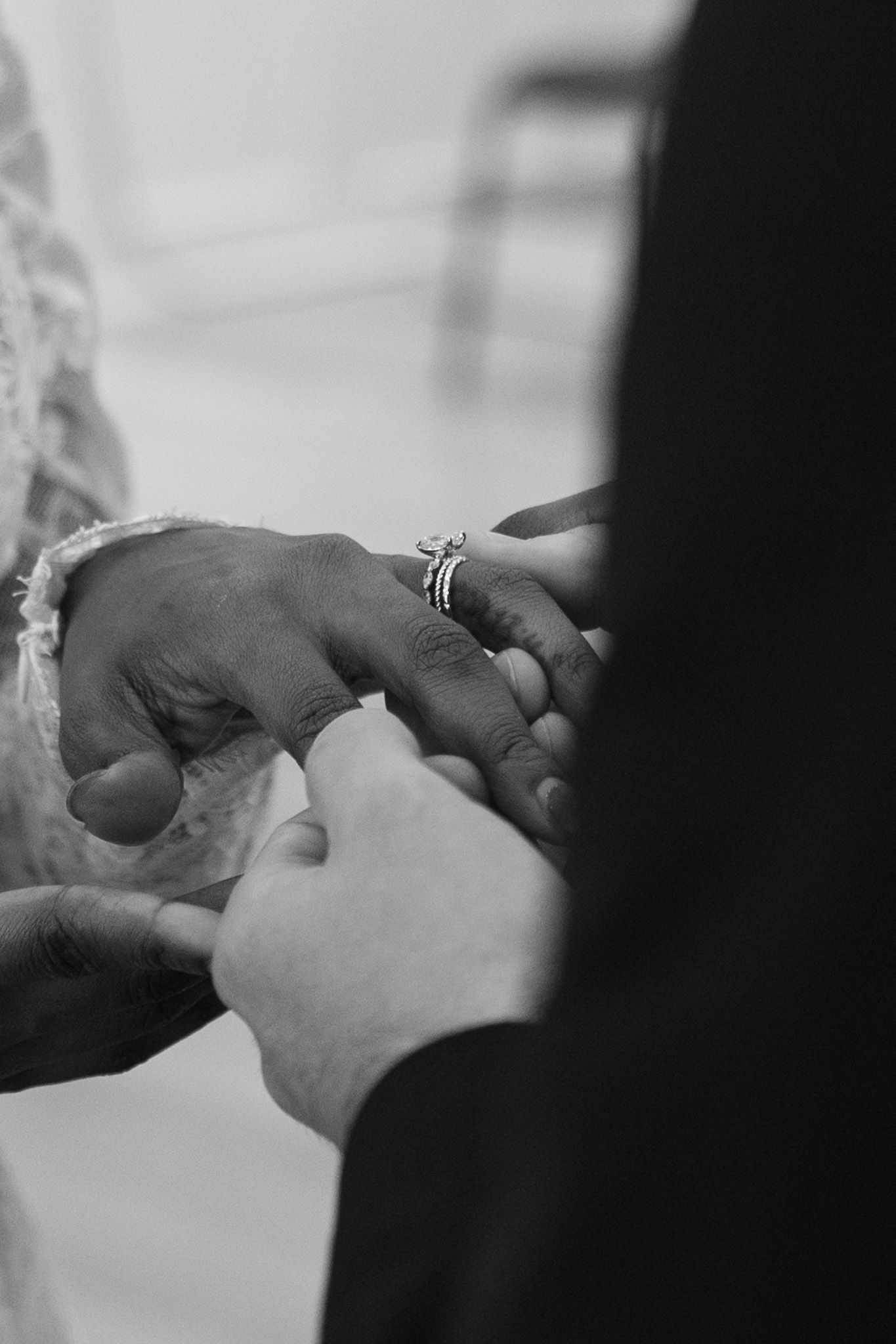 Black and white photo of a groom placing a diamond wedding ring on the bride's finger during a wedding ceremony.