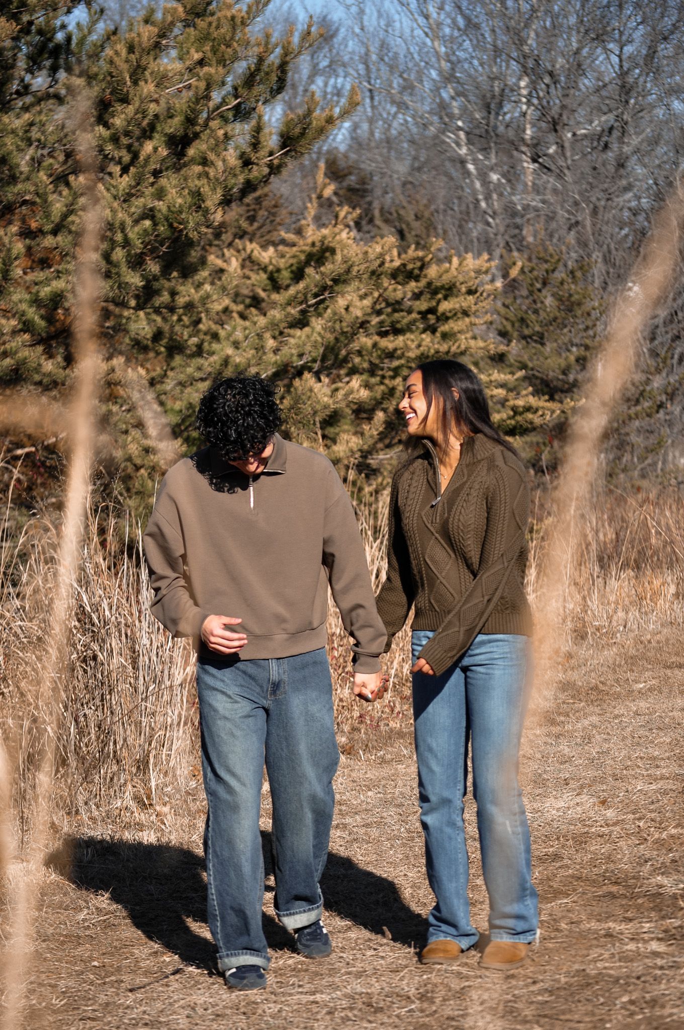 A couple holding hands and laughing while walking on a dirt path surrounded by trees in autumn.