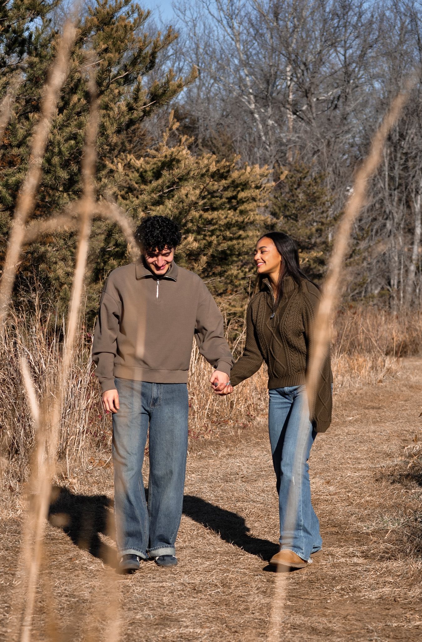 A couple holds hands and smiles while walking on a nature trail surrounded by dry grass and evergreen trees.
