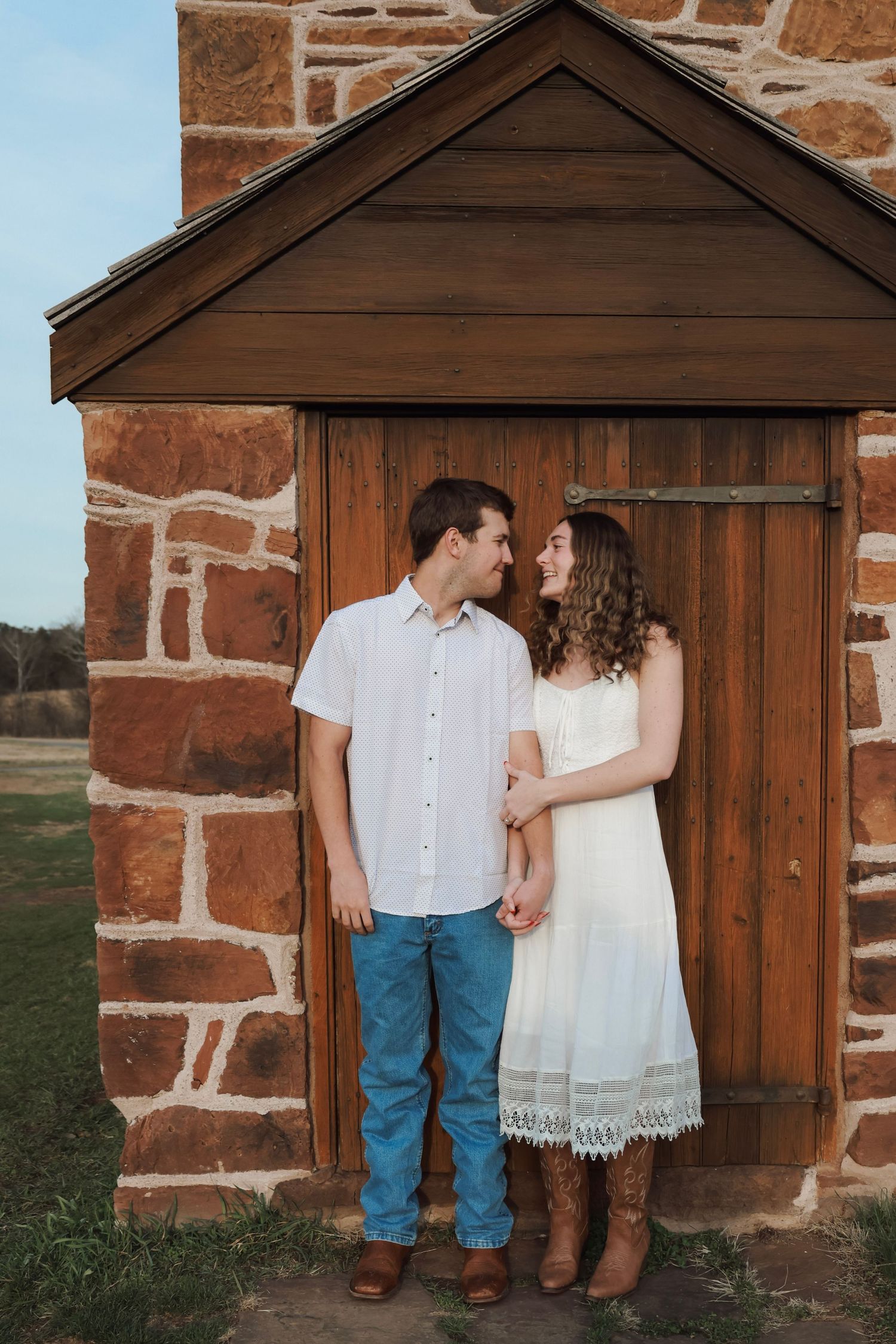 Couple in white dress and jeans holding hands, standing before rustic stone building with wooden door.