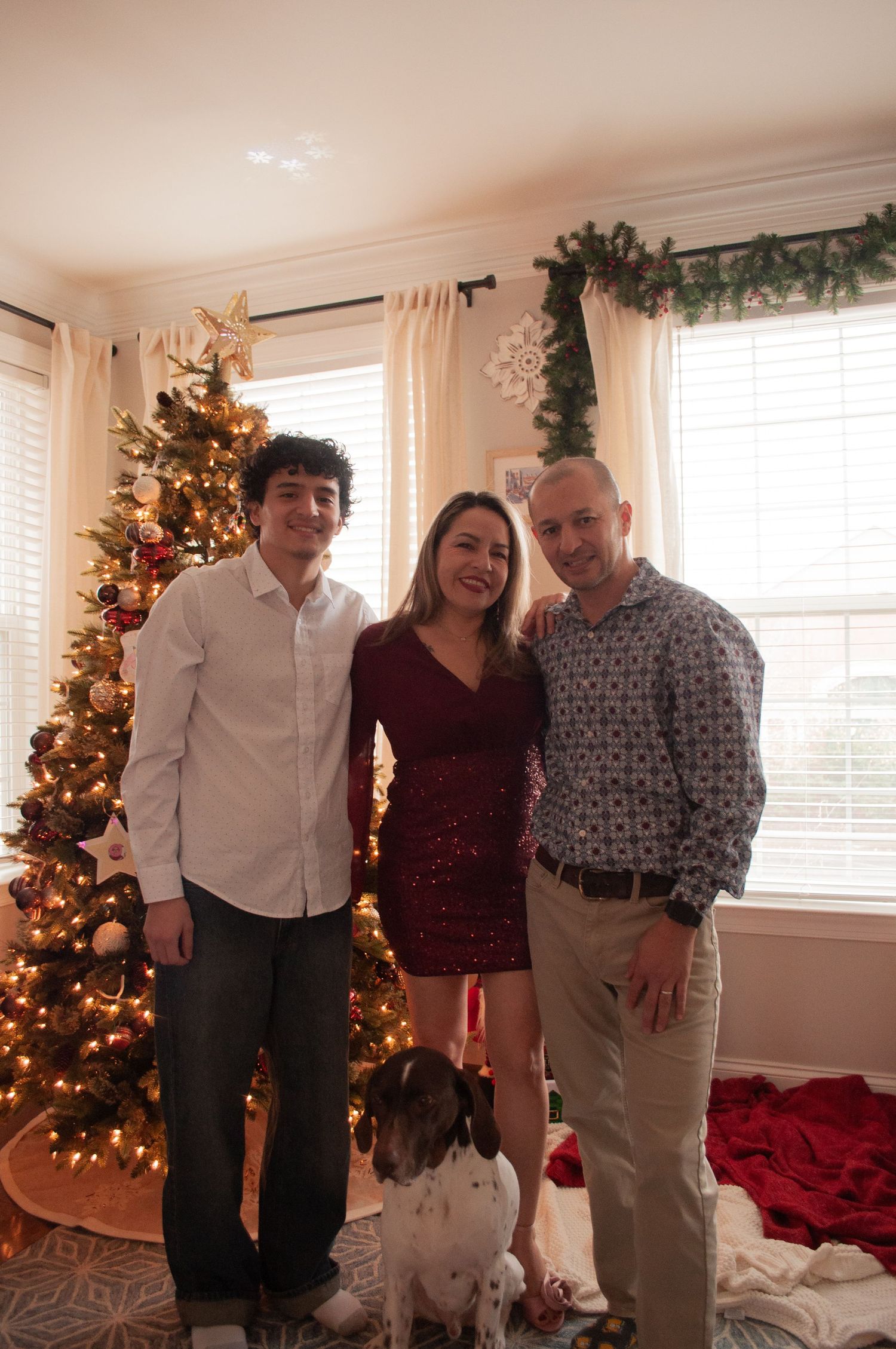 Family of three posing with their dog in front of a decorated Christmas tree in a festive holiday home.