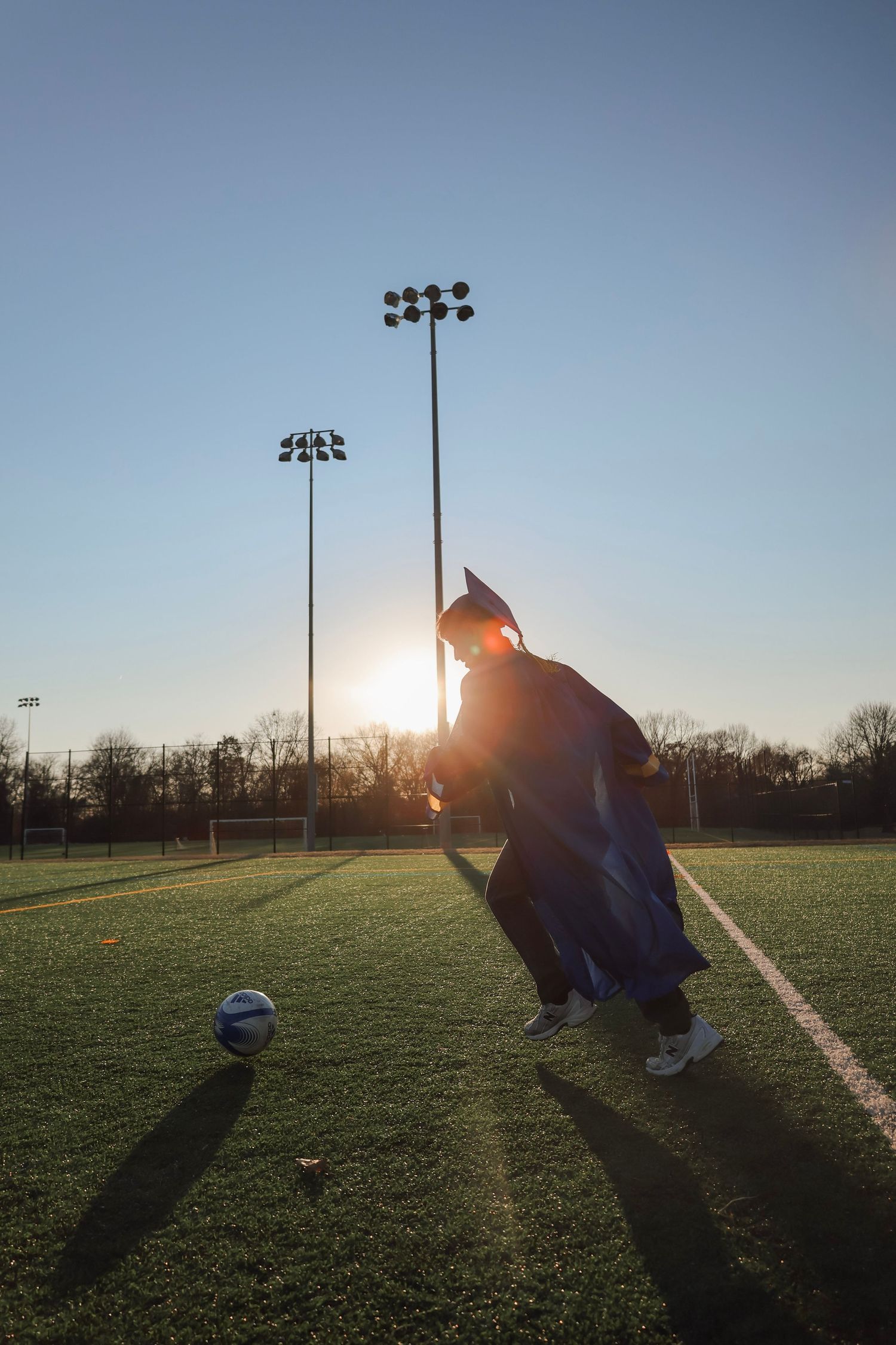 Graduate in cap and gown kicking soccer ball on green field at sunset with stadium lights in background.