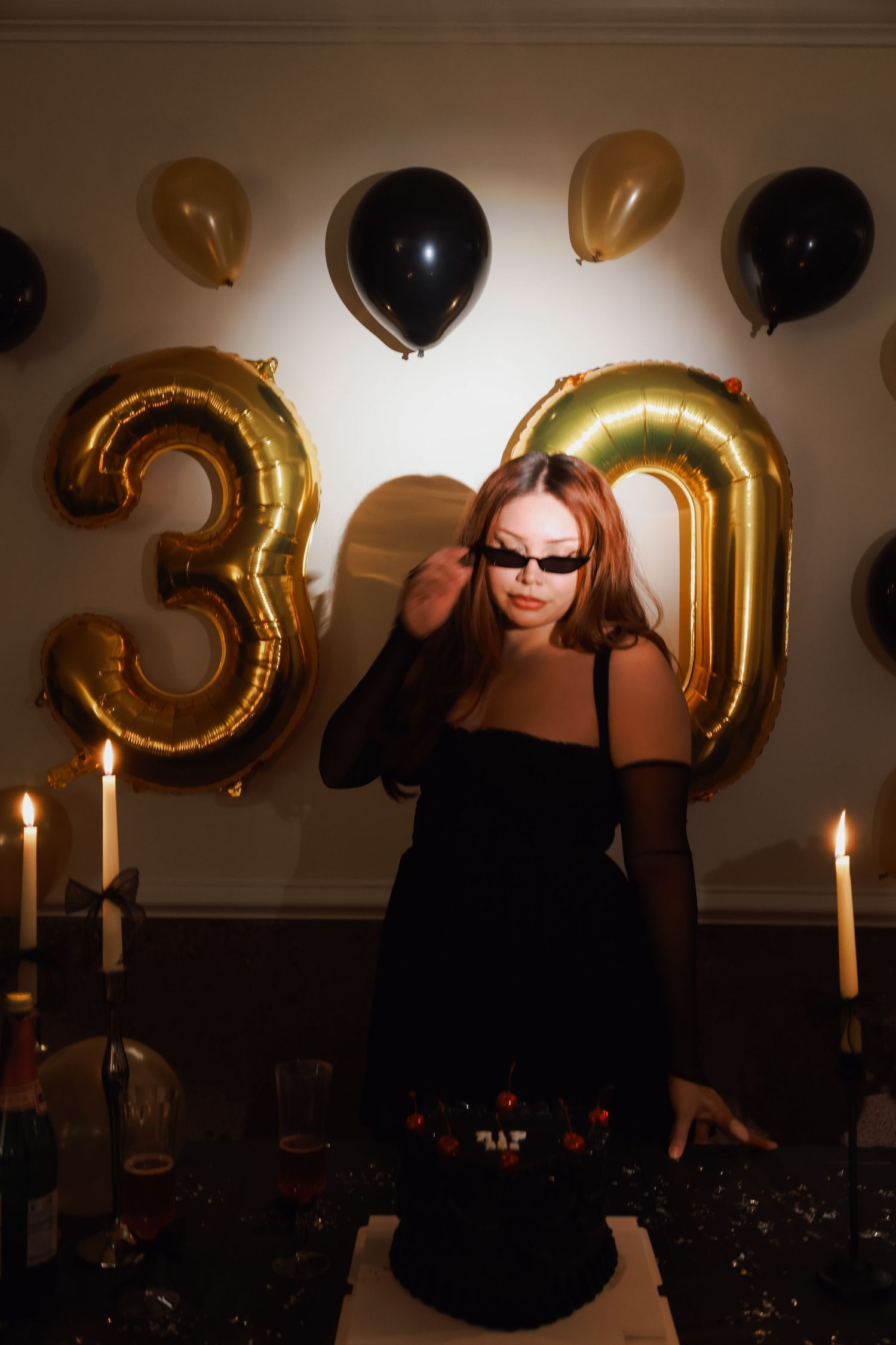Woman in black dress celebrating 30th birthday with gold balloons, candles, and cake at elegant party.