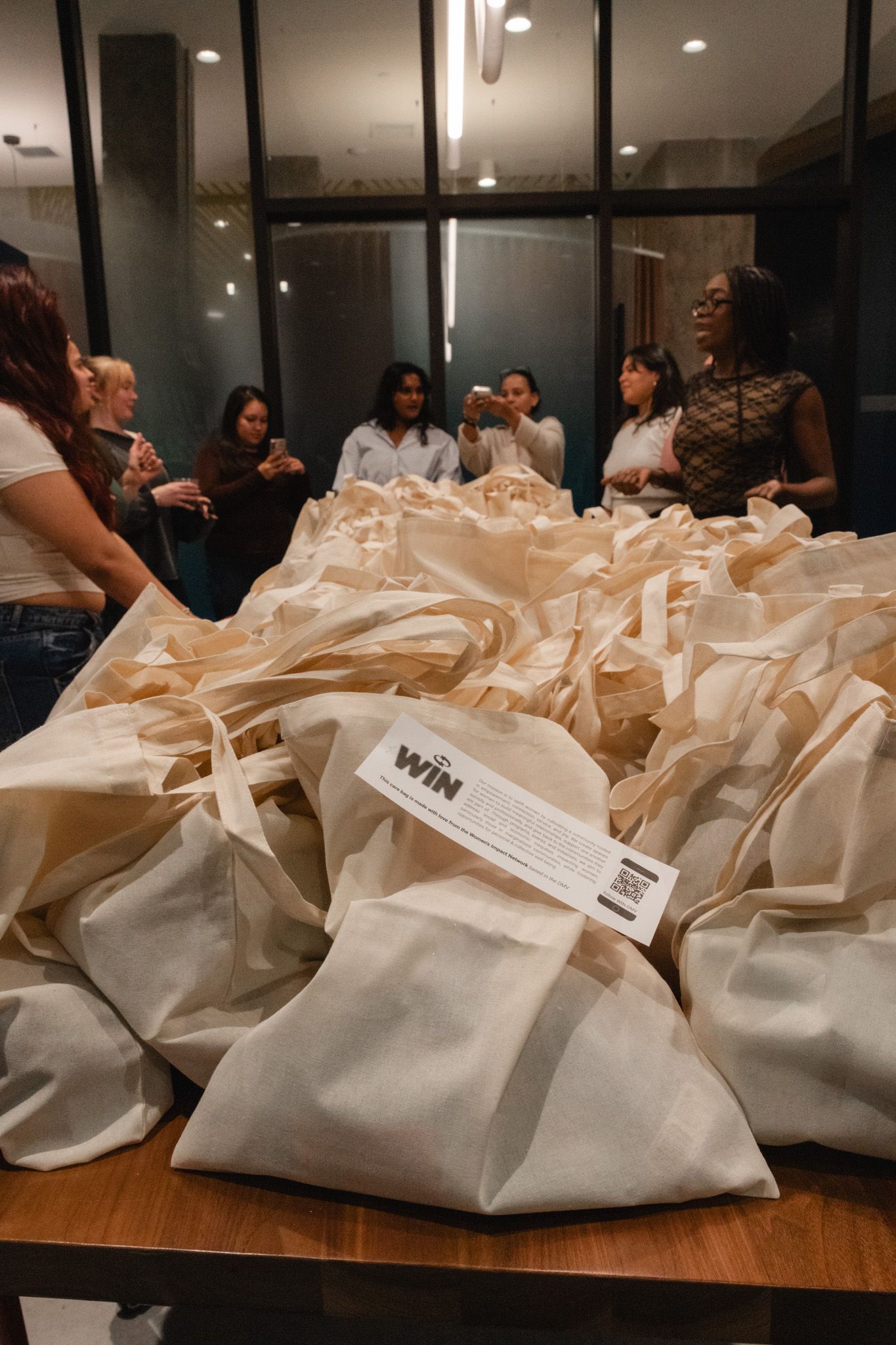 A table filled with canvas tote bags labeled WIN at an indoor event with a group of women gathered in the background.