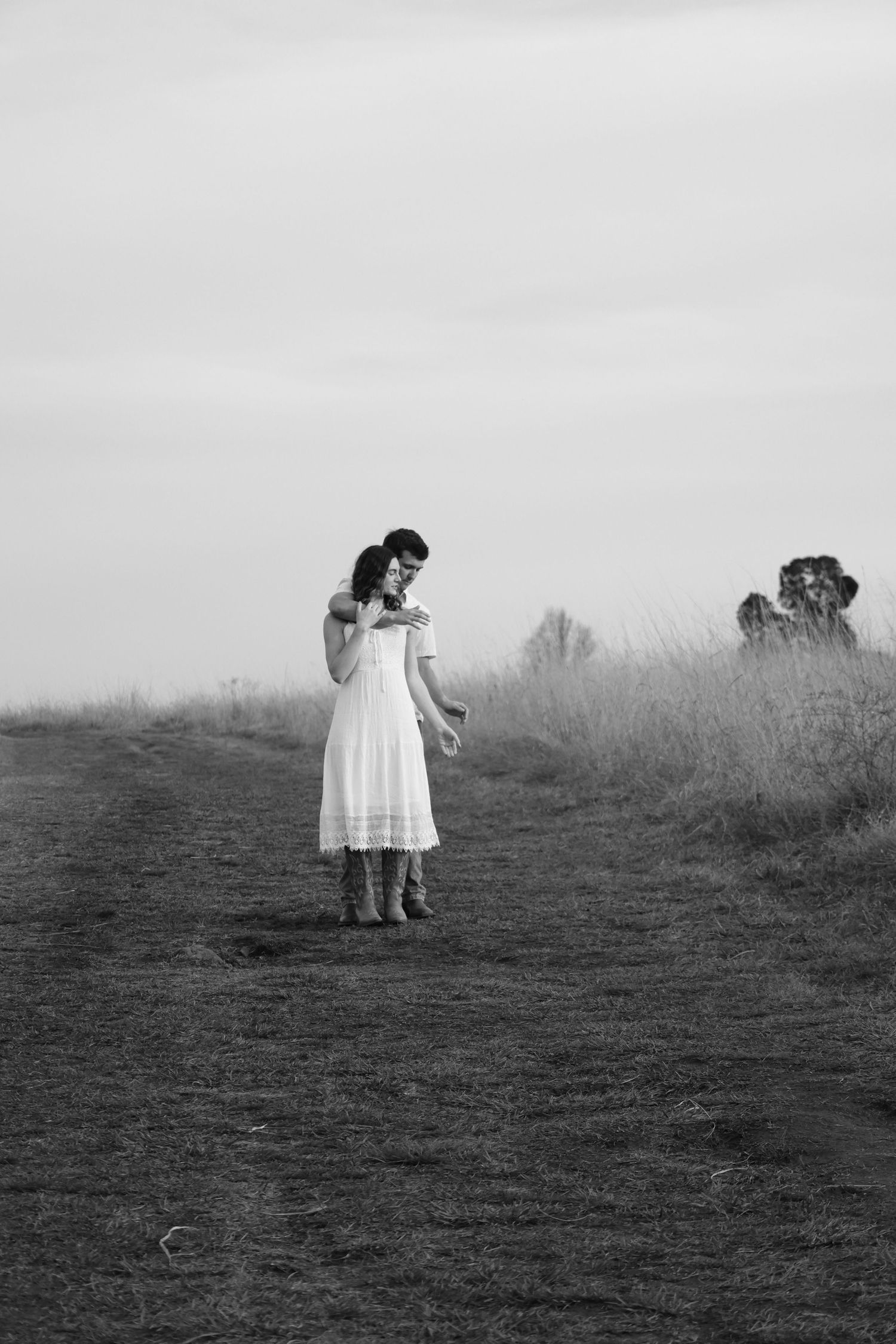 Black and white photo of a couple embracing on a rural dirt path surrounded by tall grass under a cloudy sky.