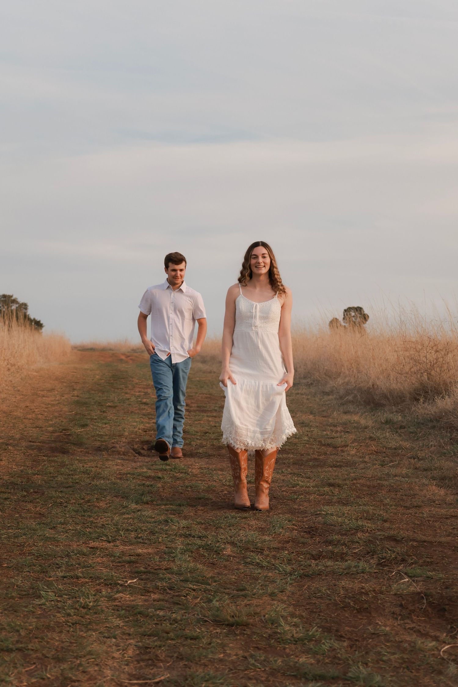 A couple walks along a rural dirt path, woman in white dress and cowboy boots, man in jeans and white shirt, golden fields.