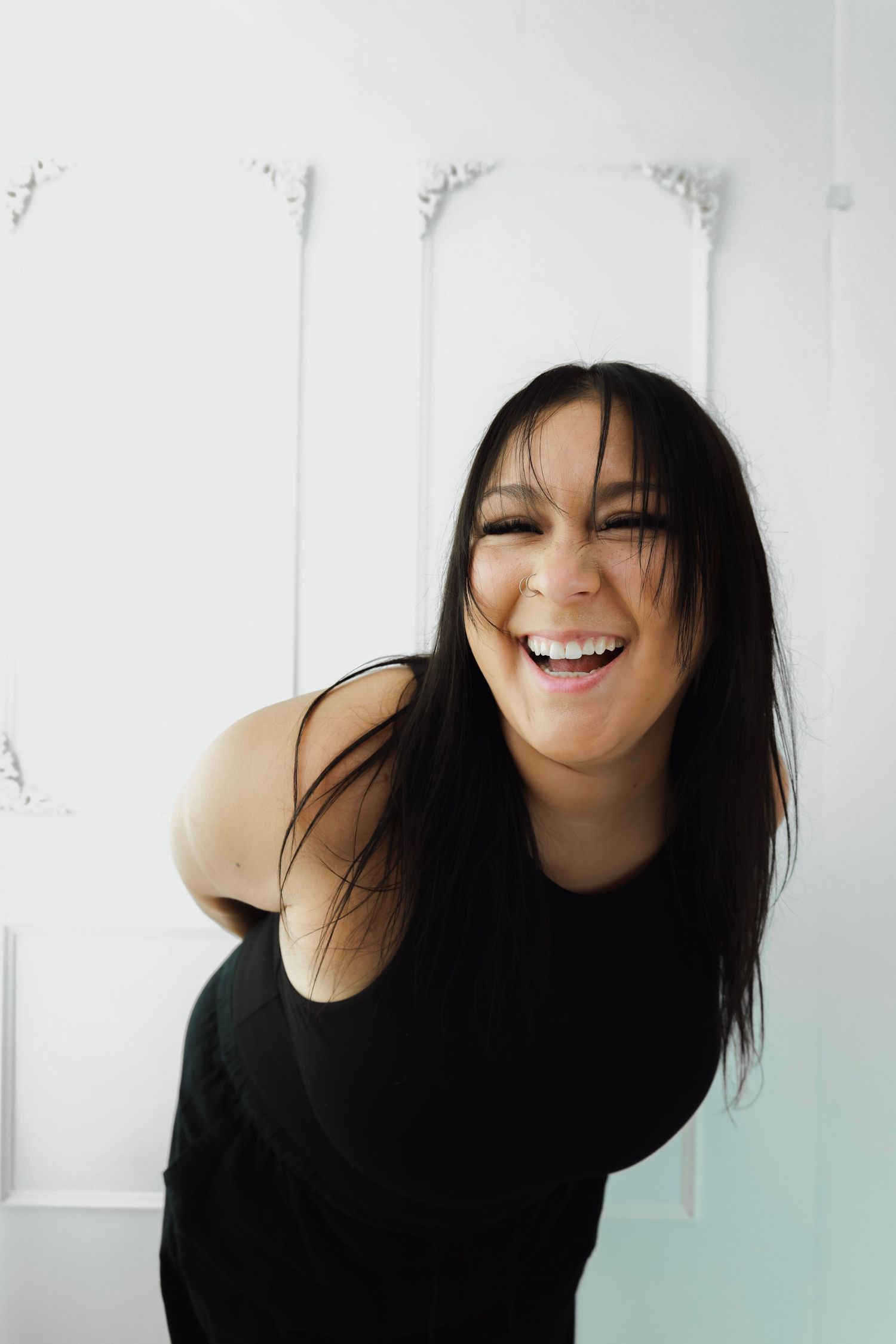 A young woman with long dark hair laughs joyfully, wearing a black top against a white decorative wall in a studio.