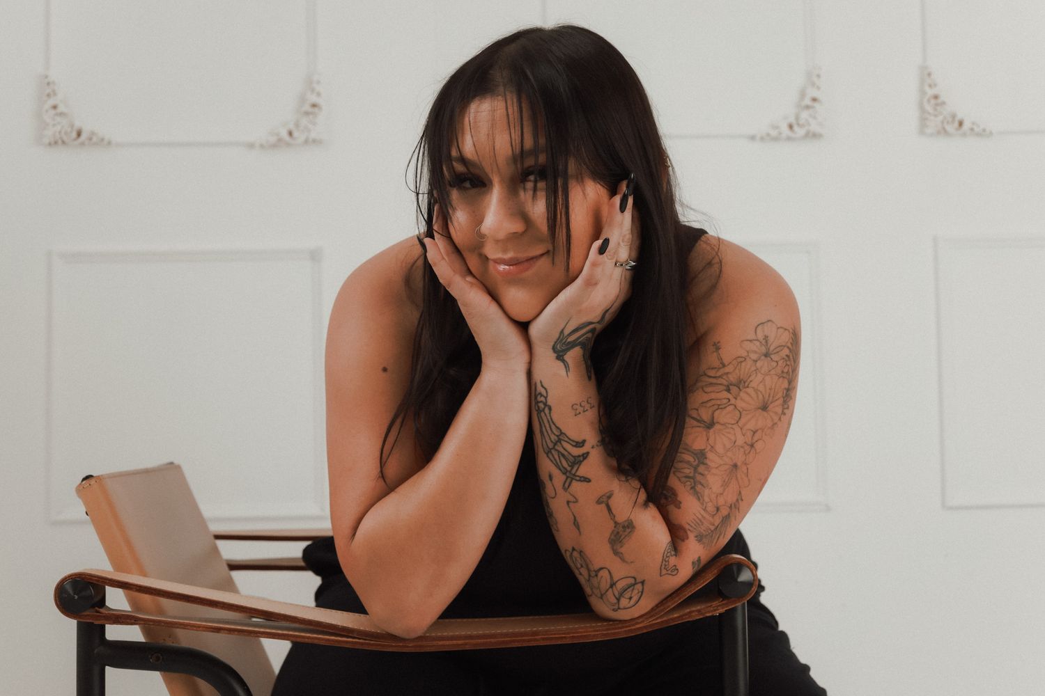 Tattooed woman with long dark hair resting chin on hands at wooden desk in white studio.