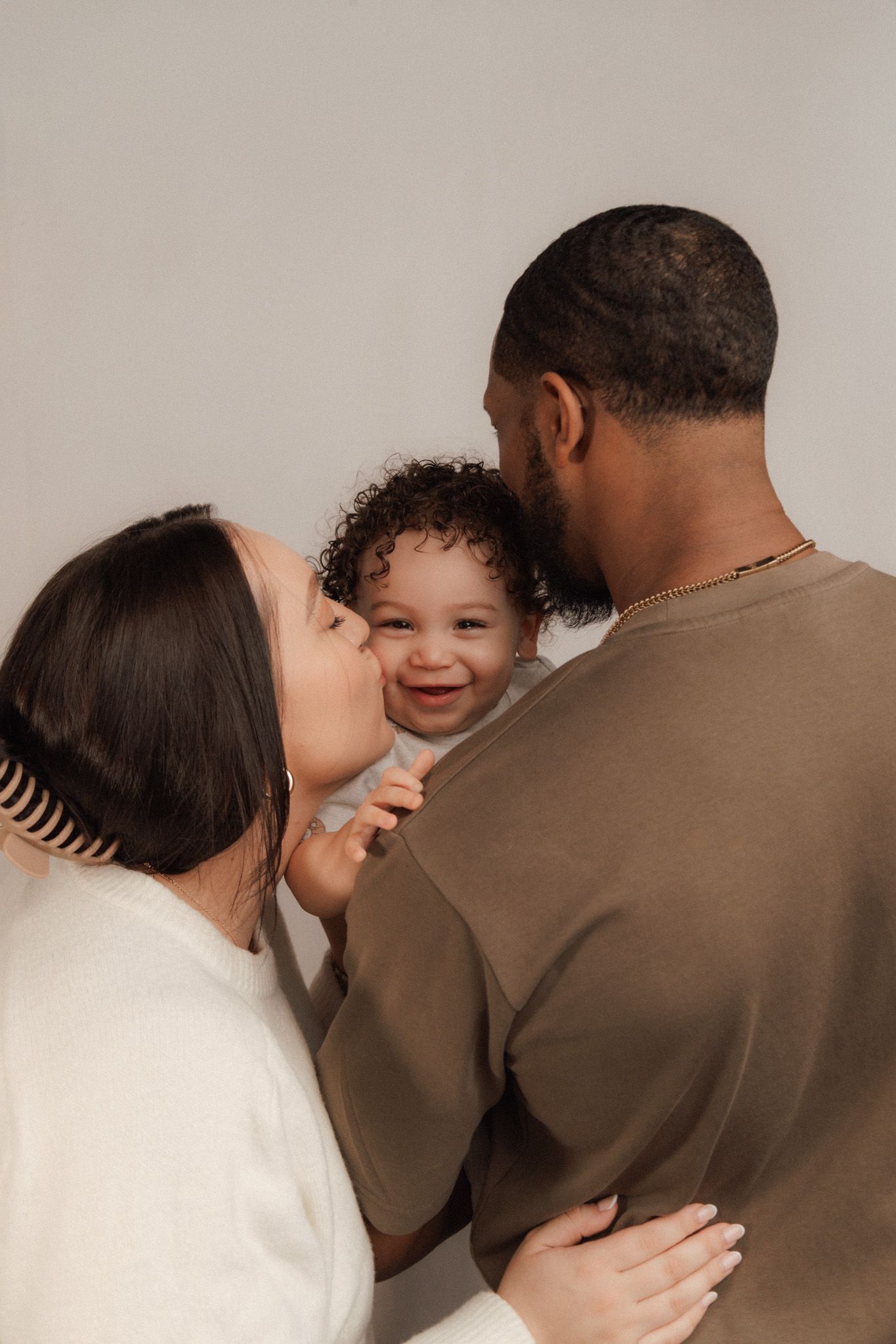 A joyful baby smiles as mom kisses their cheek while dad holds them in a warm family portrait.