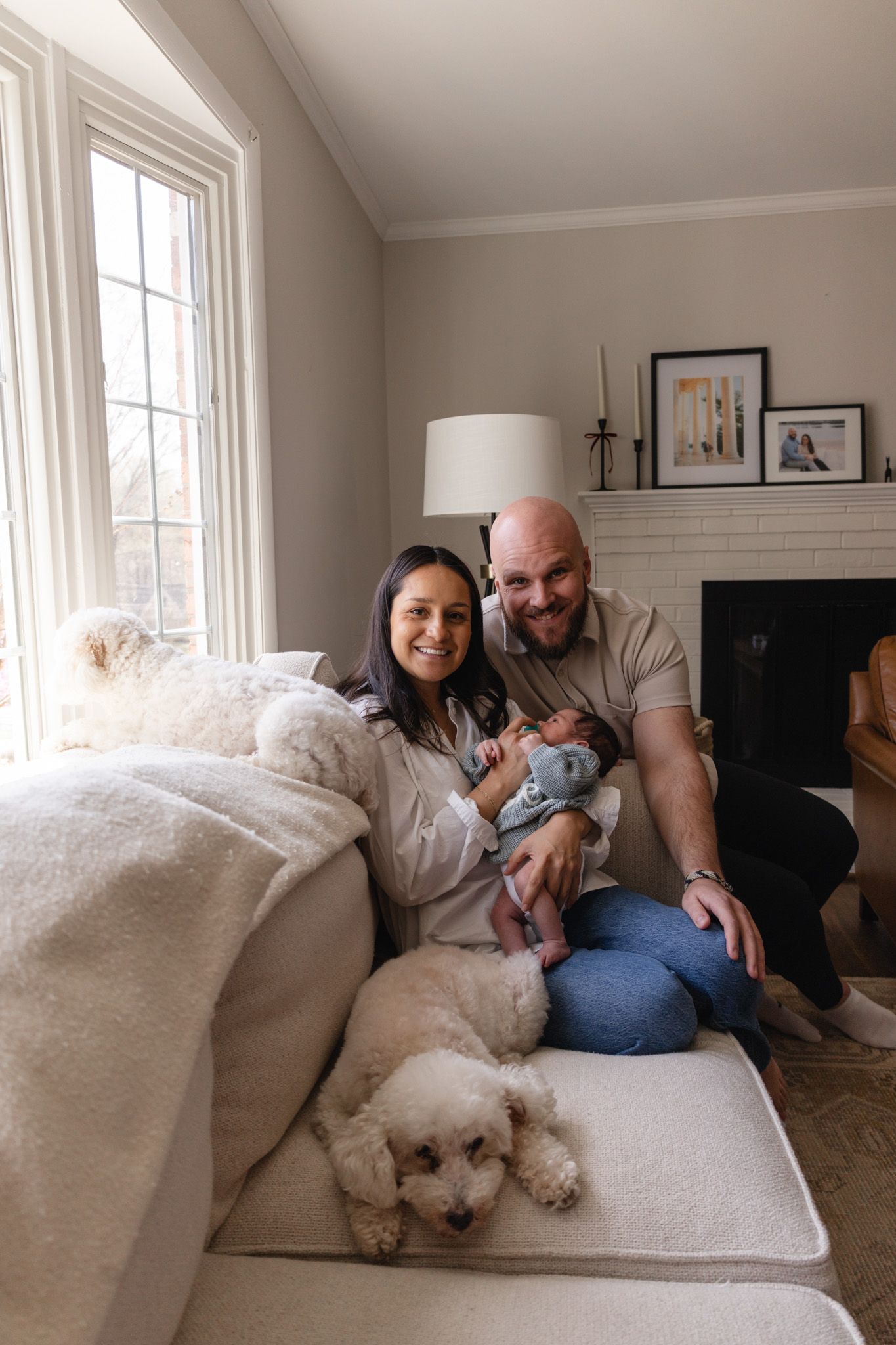 A smiling couple sits on a couch with their newborn baby and two fluffy white dogs in a cozy living room.