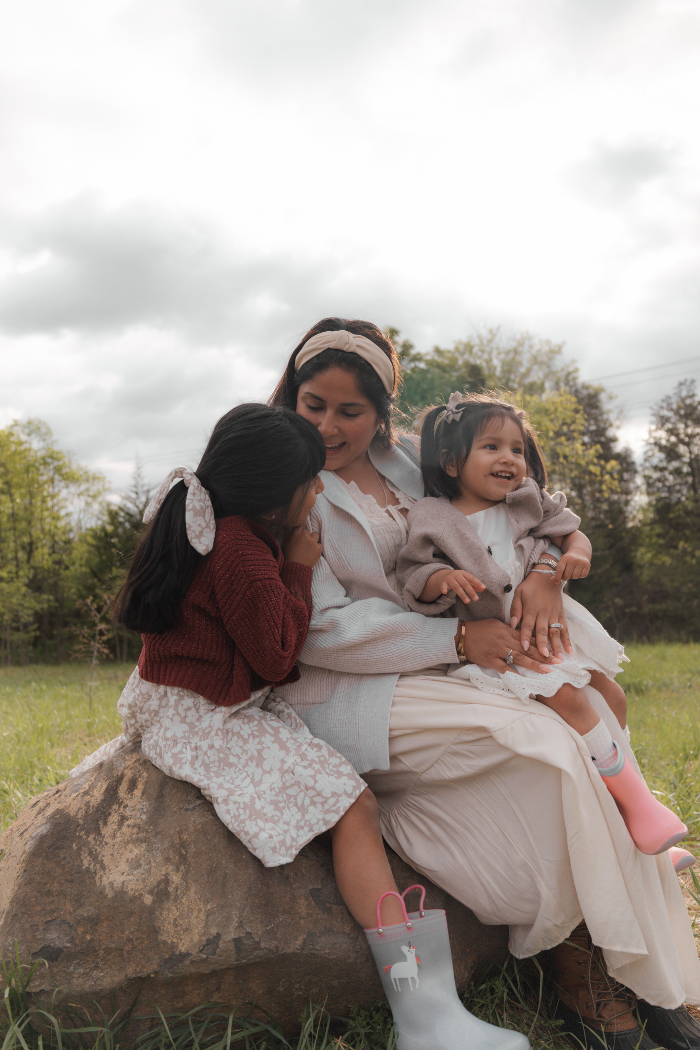 A mother sits on a rock in a field, smiling as her two young daughters cuddle close in matching floral outfits.
