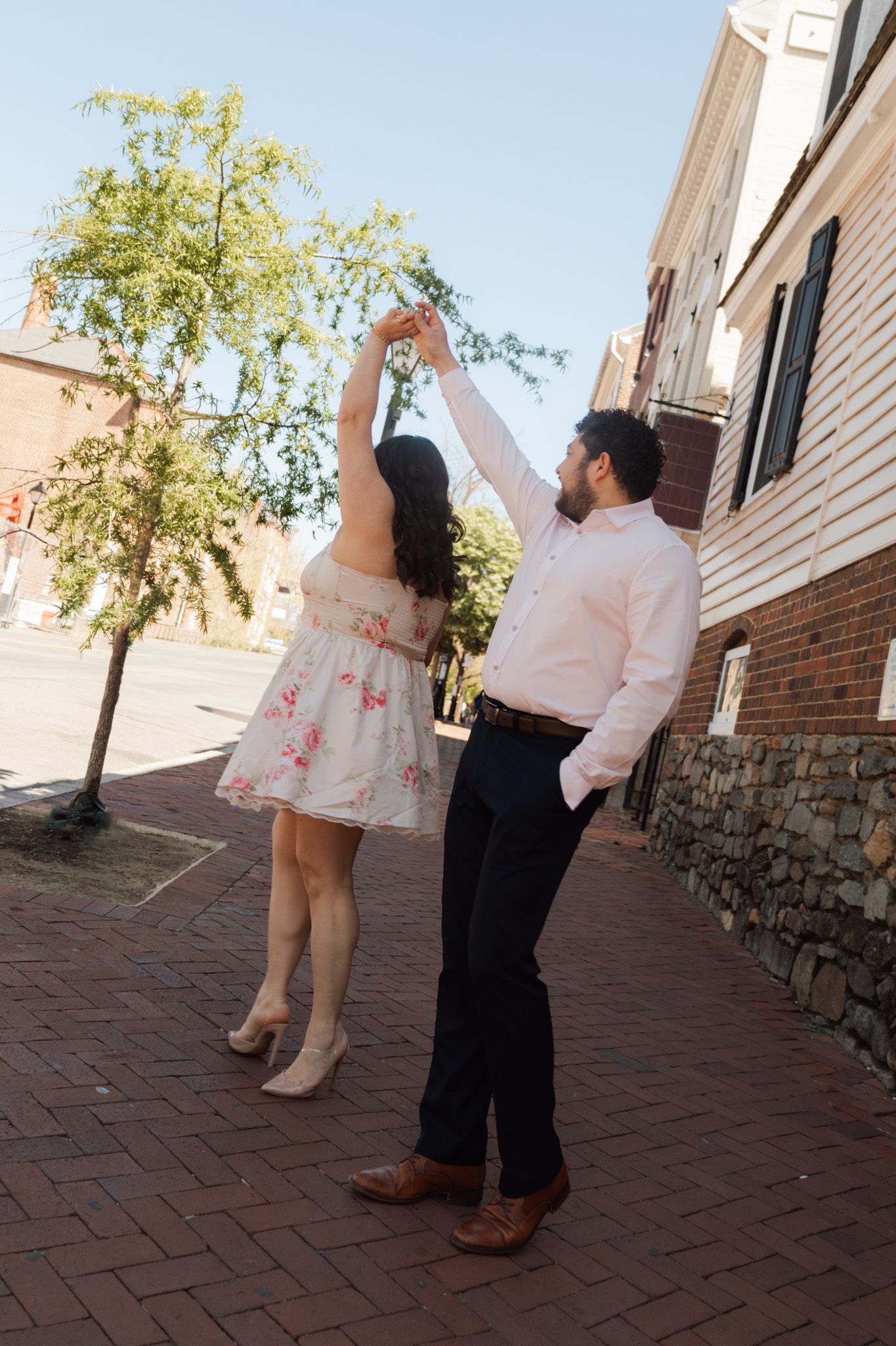 Couple dancing on a brick sidewalk in a charming town, woman in floral dress being twirled by man in white shirt.