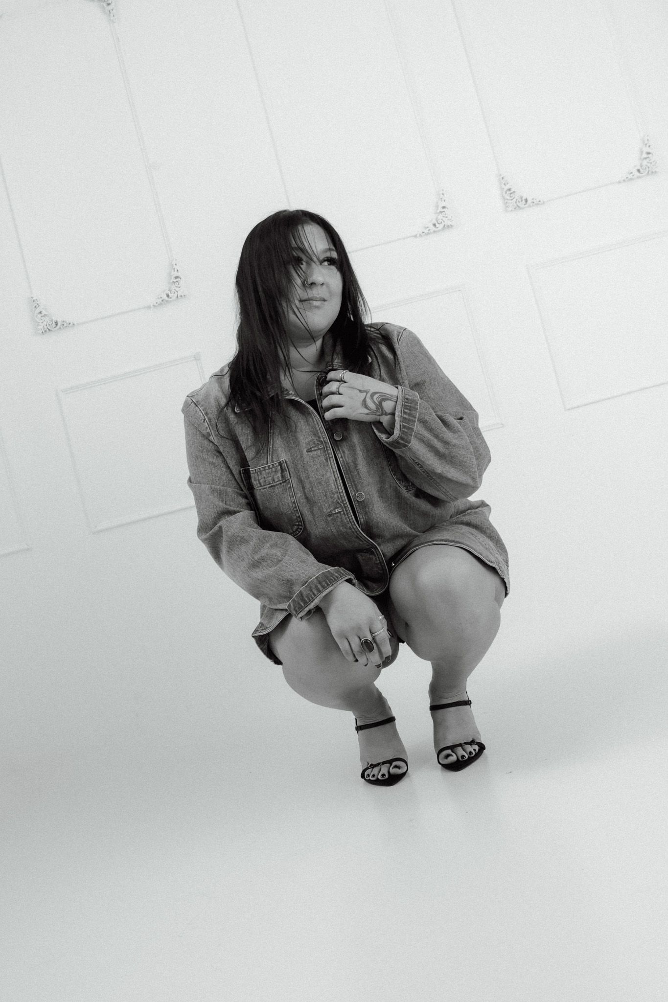 Black and white photo of a woman in a denim outfit crouching gracefully against a white decorative wall.