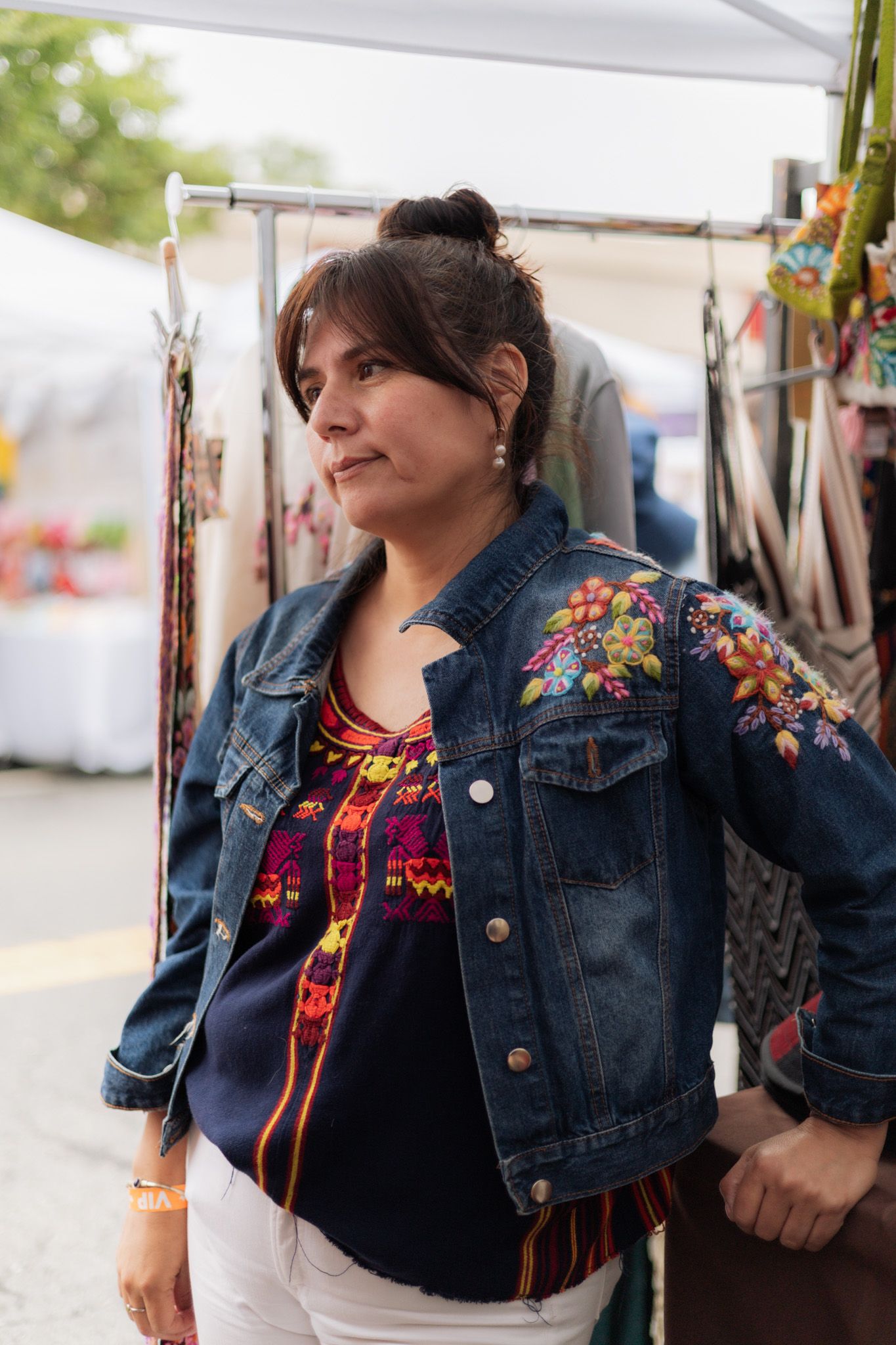 Woman in embroidered denim jacket at outdoor market stall, wearing colorful traditional blouse and white pants.
