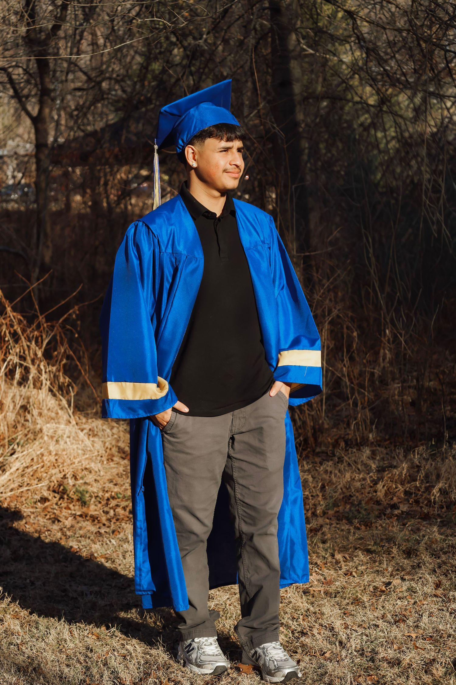 Young man in blue graduation cap and gown posing outdoors among bare trees on a sunny day.